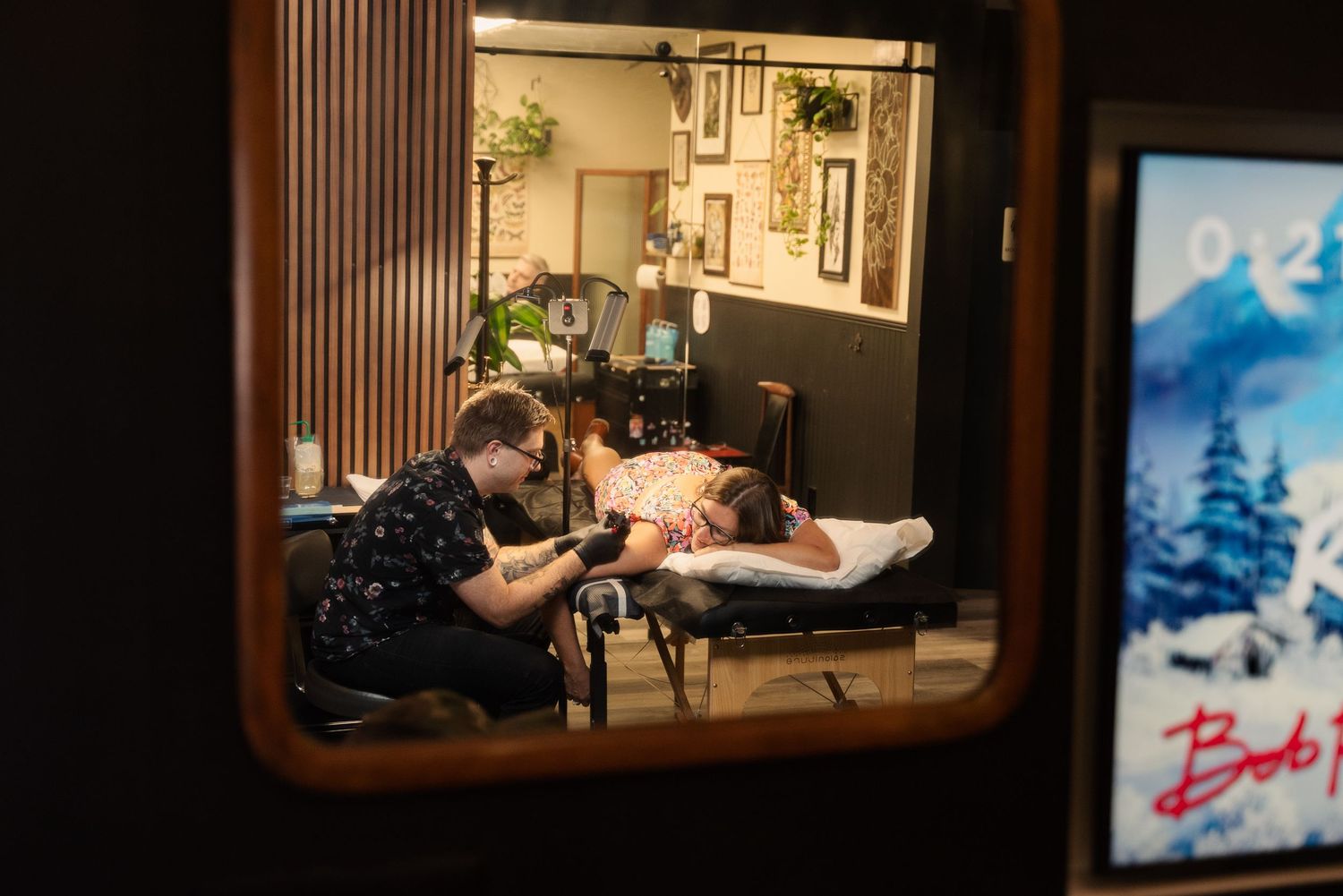 View through studio windows showing tattoo session in progress with mountain artwork on walls.