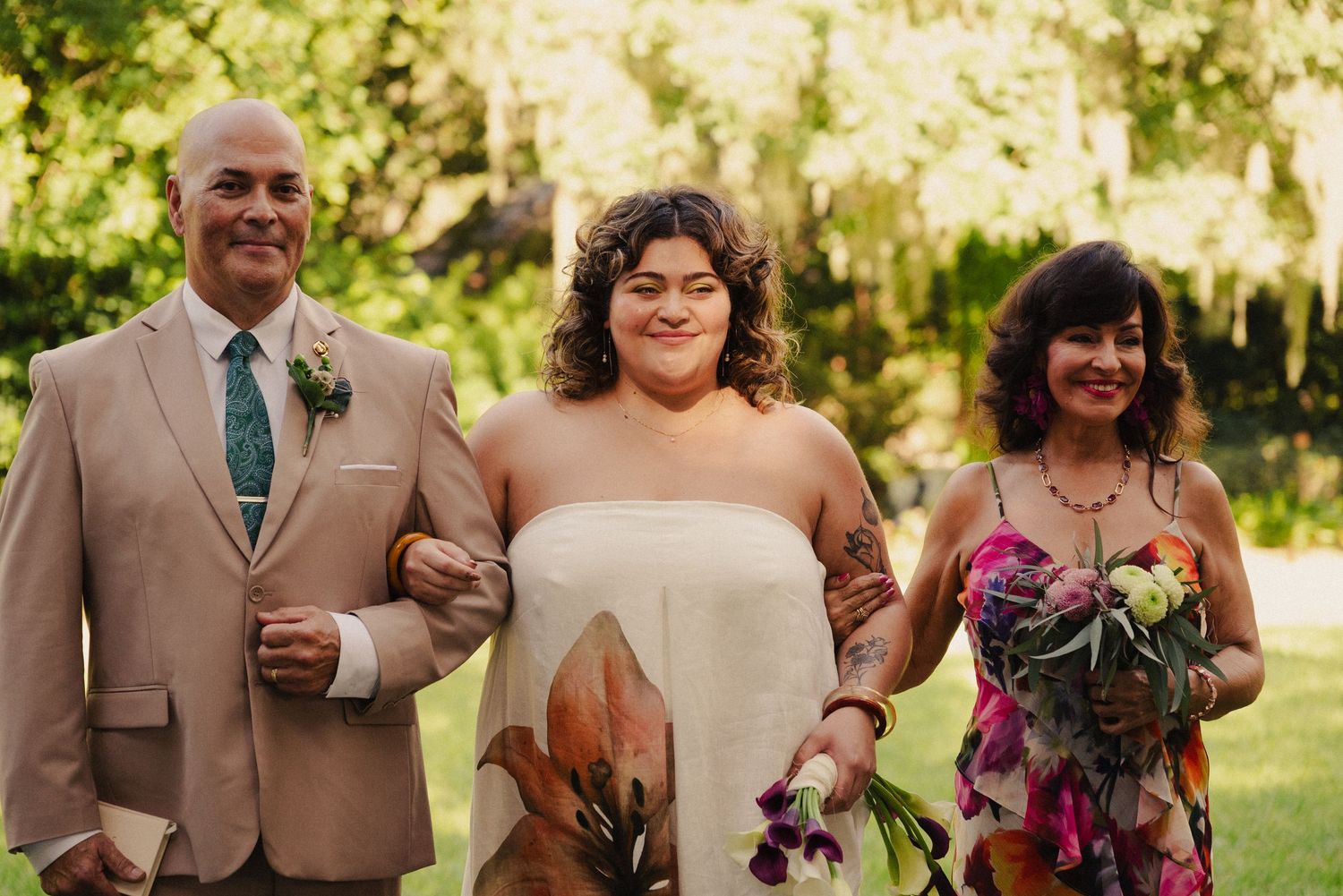 Wedding party members stand together outdoors wearing brown suits and holding flowers.