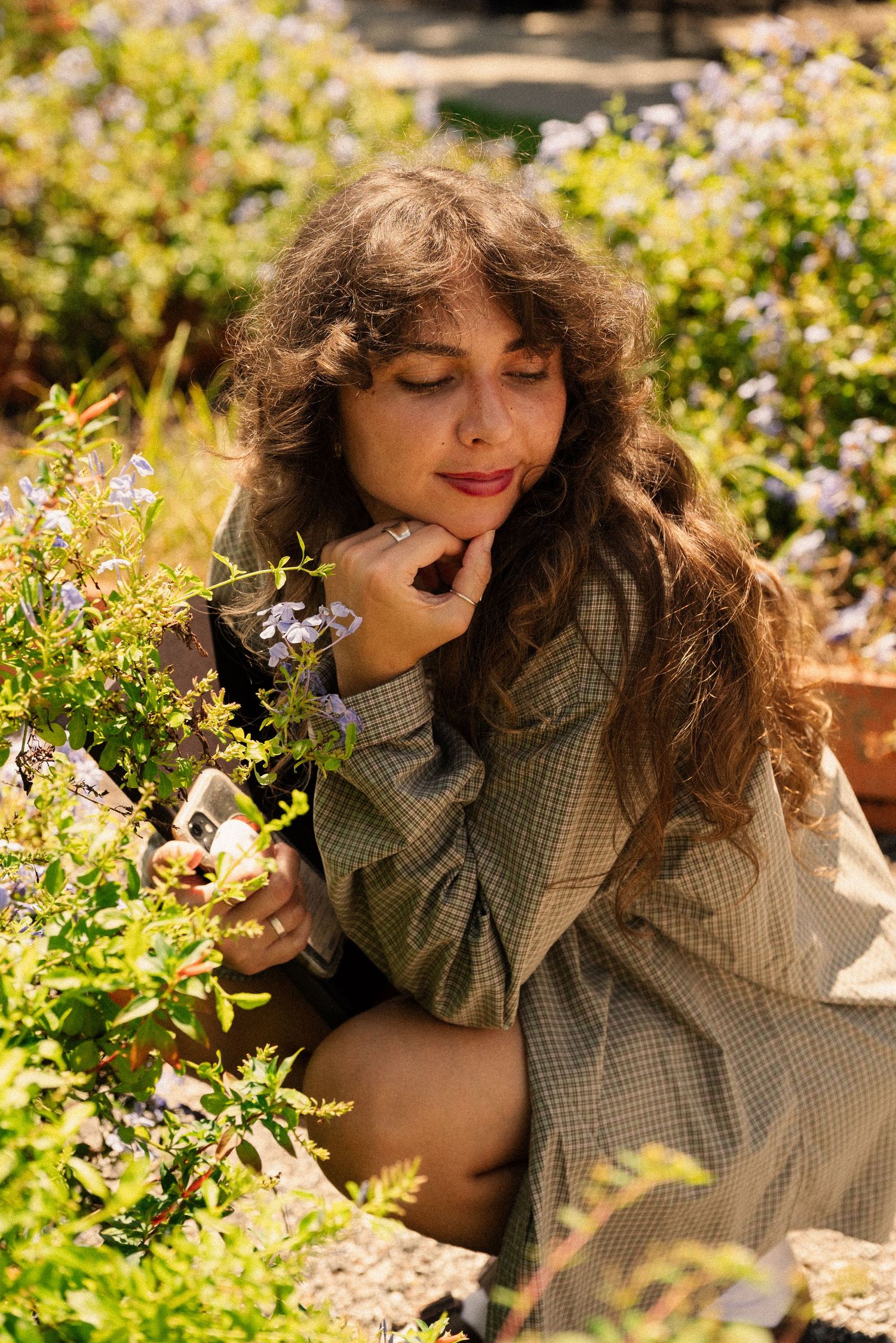 Woman in gingham shirt poses artistically among blooming flowers in a sunny garden setting.