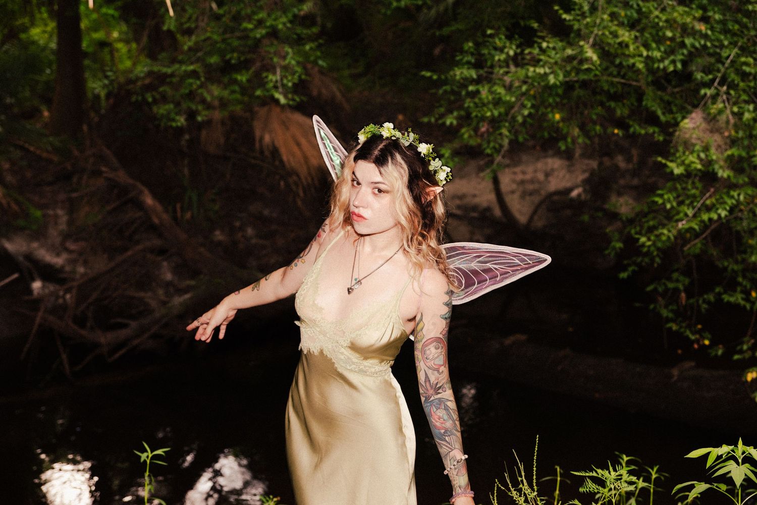 Woodland fairy wearing flower crown and silk dress poses near dark water at dusk.