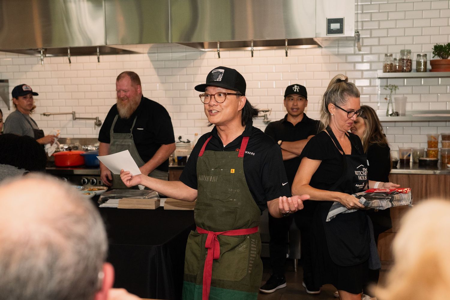 Kitchen staff in green aprons working in bright restaurant kitchen with white subway tile walls.
