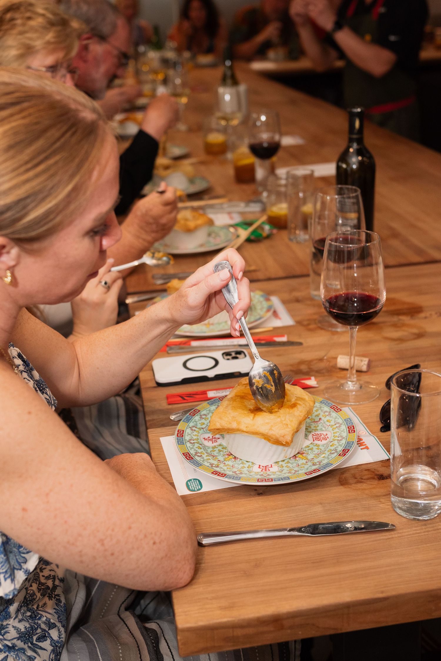 Diners enjoying meals at a wooden communal table with wine glasses and colorful plates.
