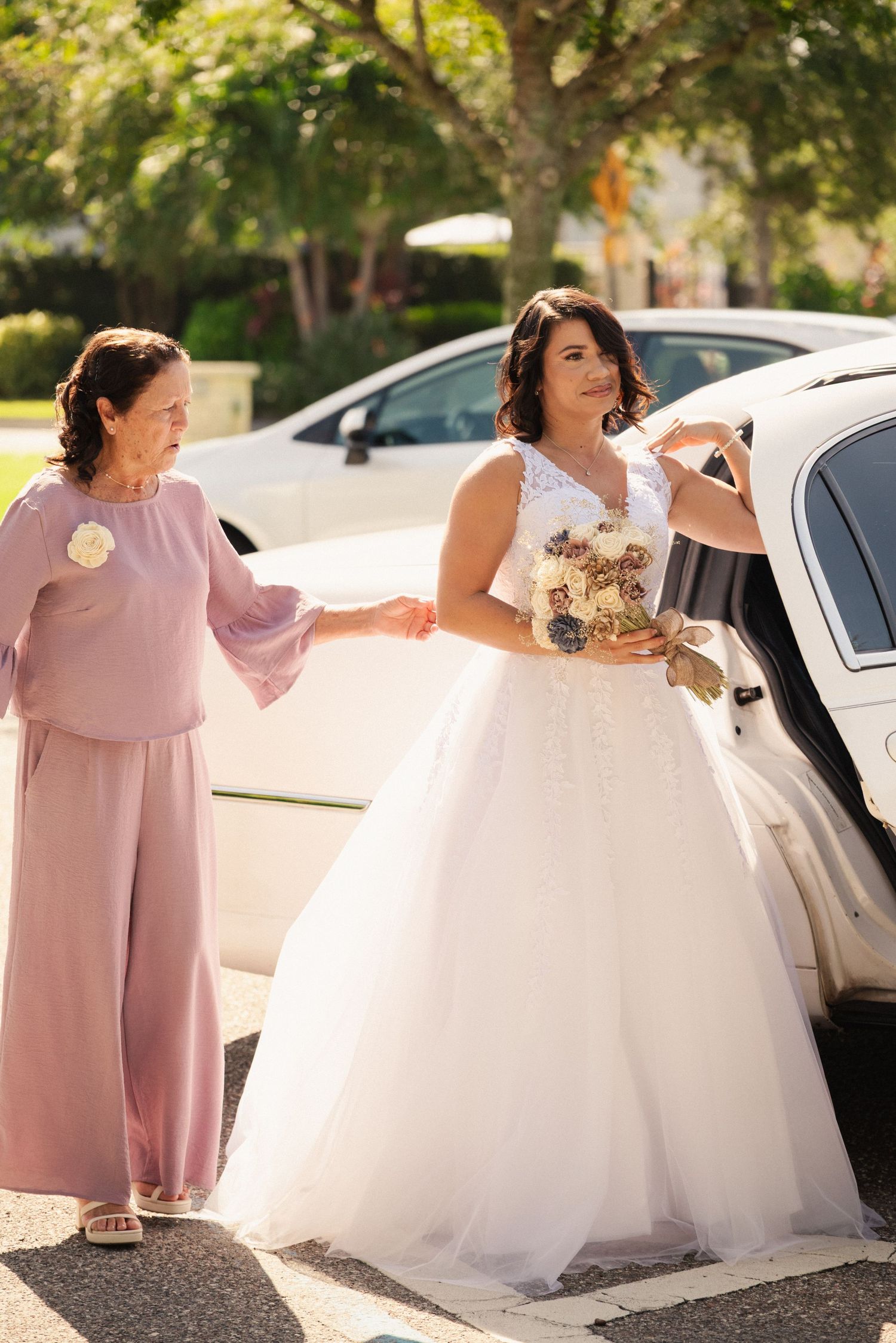 Bridesmaids in pink dresses help adjust the bride's white ballgown wedding dress in an outdoor setting.