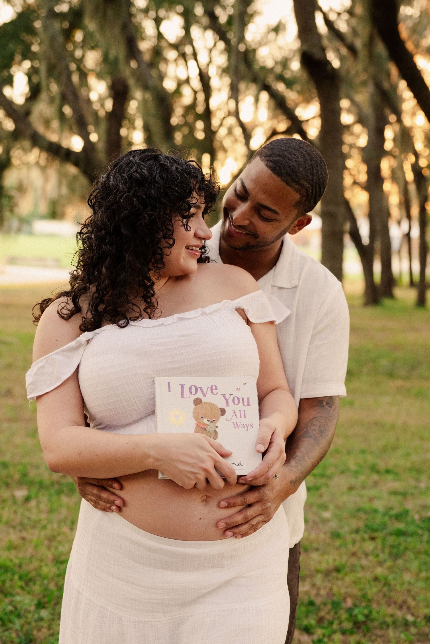 Romantic maternity photoshoot at sunset shows couple embracing in a park setting.