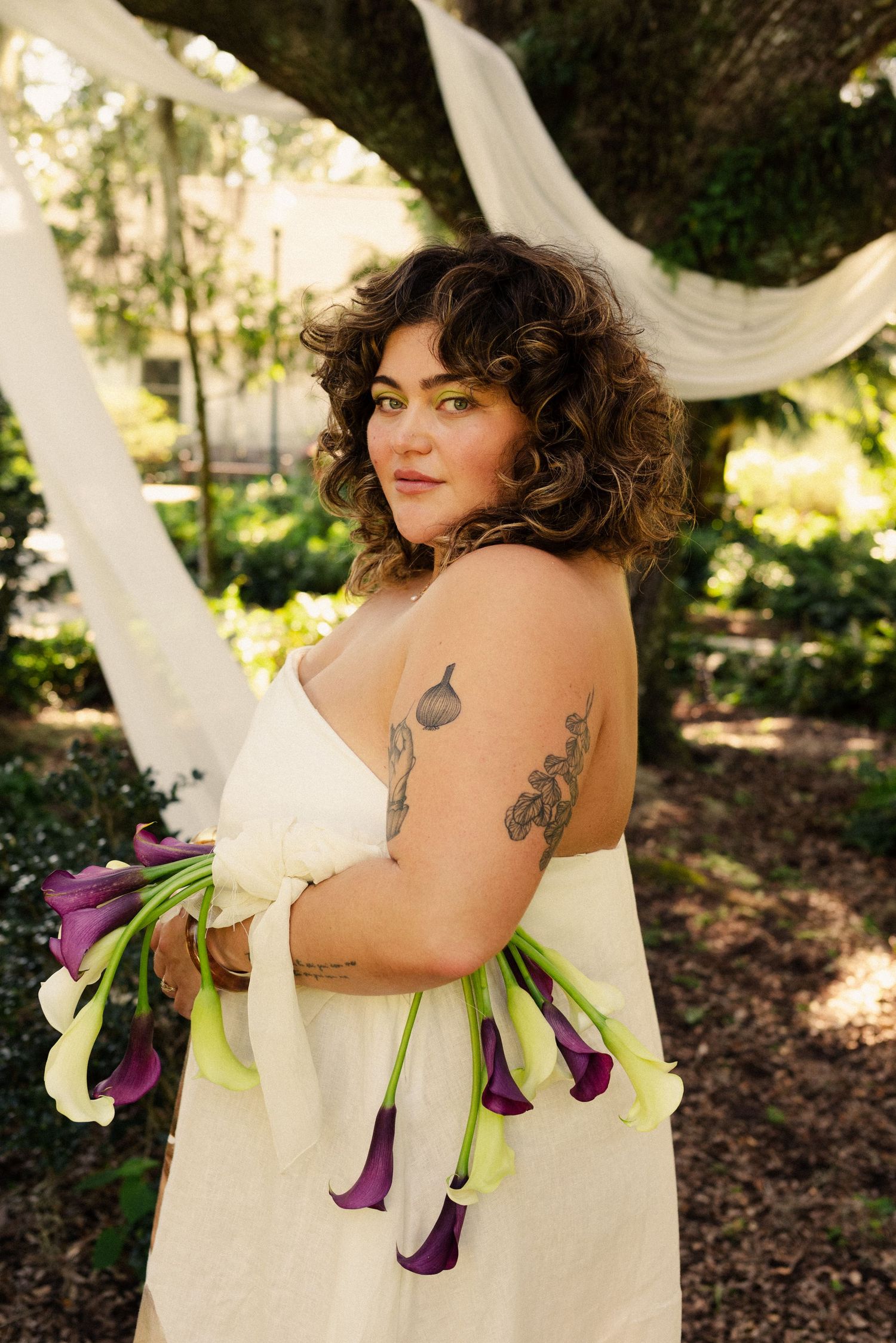A wedding dress features purple calla lilies arranged along its edges in an outdoor garden setting.
