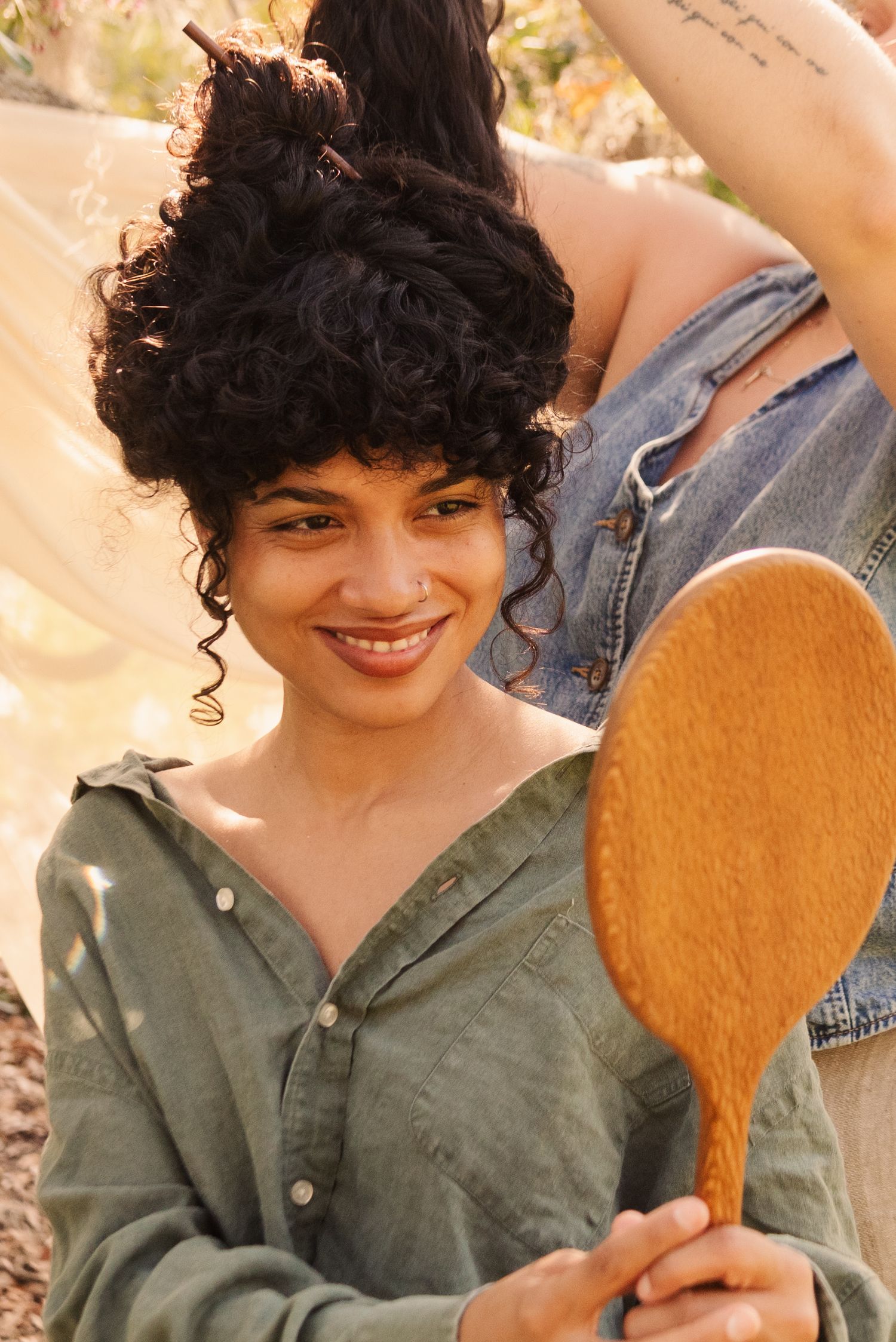 A person with curly dark hair holds up a wooden spoon while wearing a green button-up shirt in warm outdoor lighting.