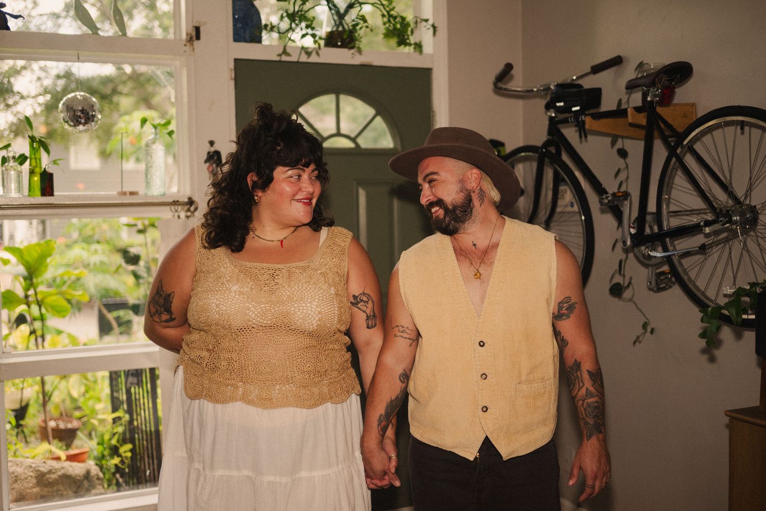 Two people in vintage-style clothing pose together in a sunlit room with bikes visible.