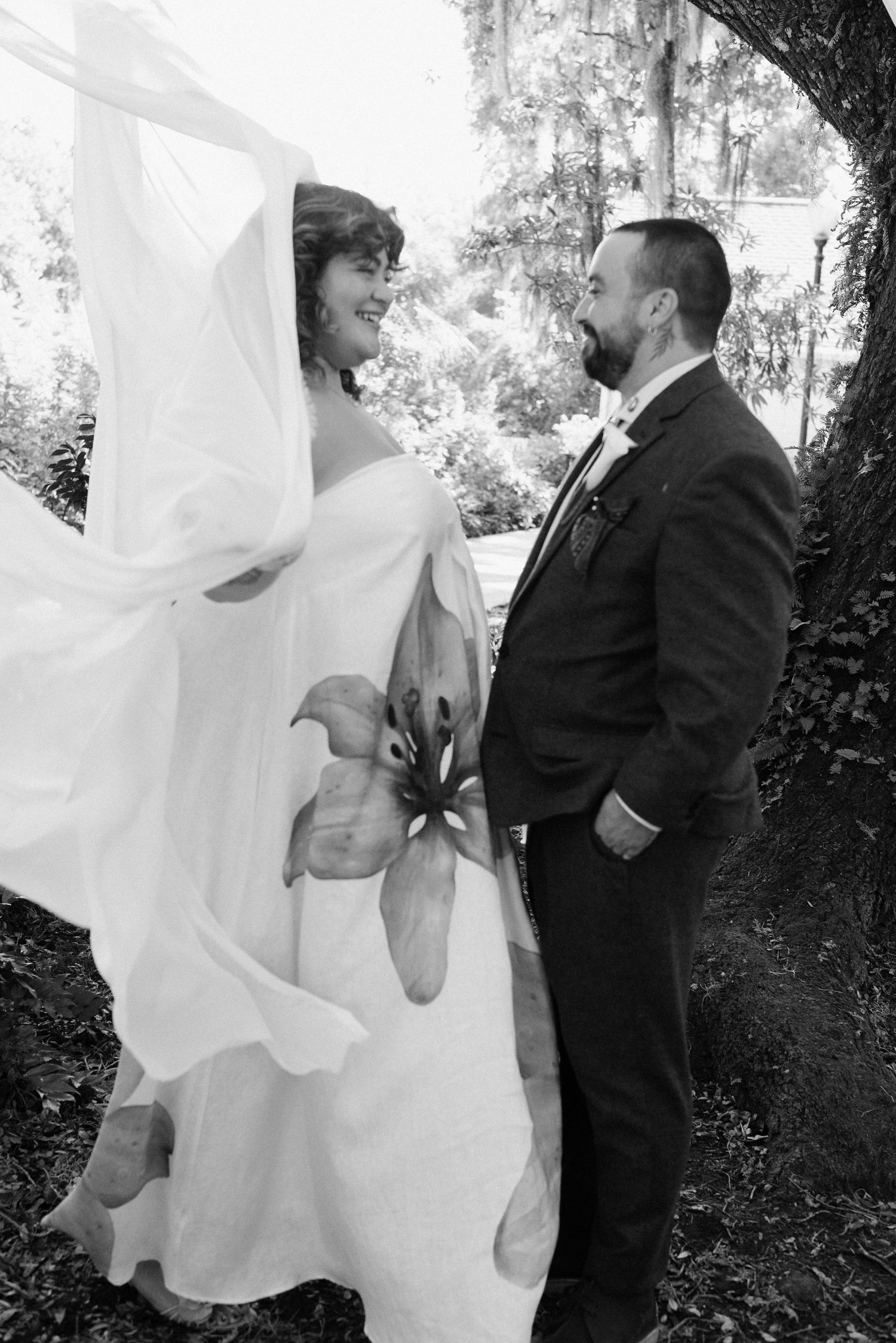 Black and white wedding portrait under trees with bride's dress flowing in the wind.