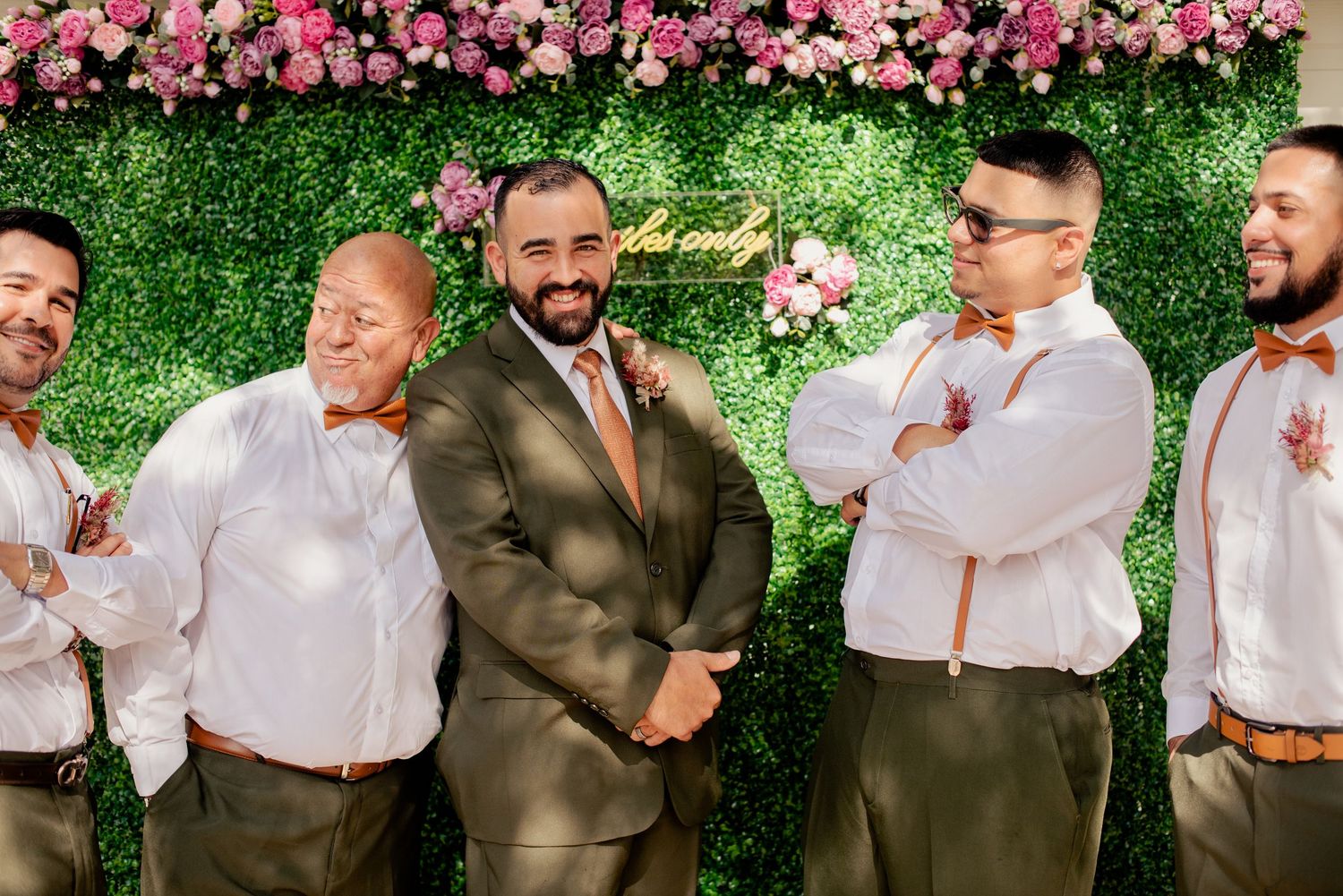 Groomsmen in brown suits and white shirts pose against floral wall backdrop.