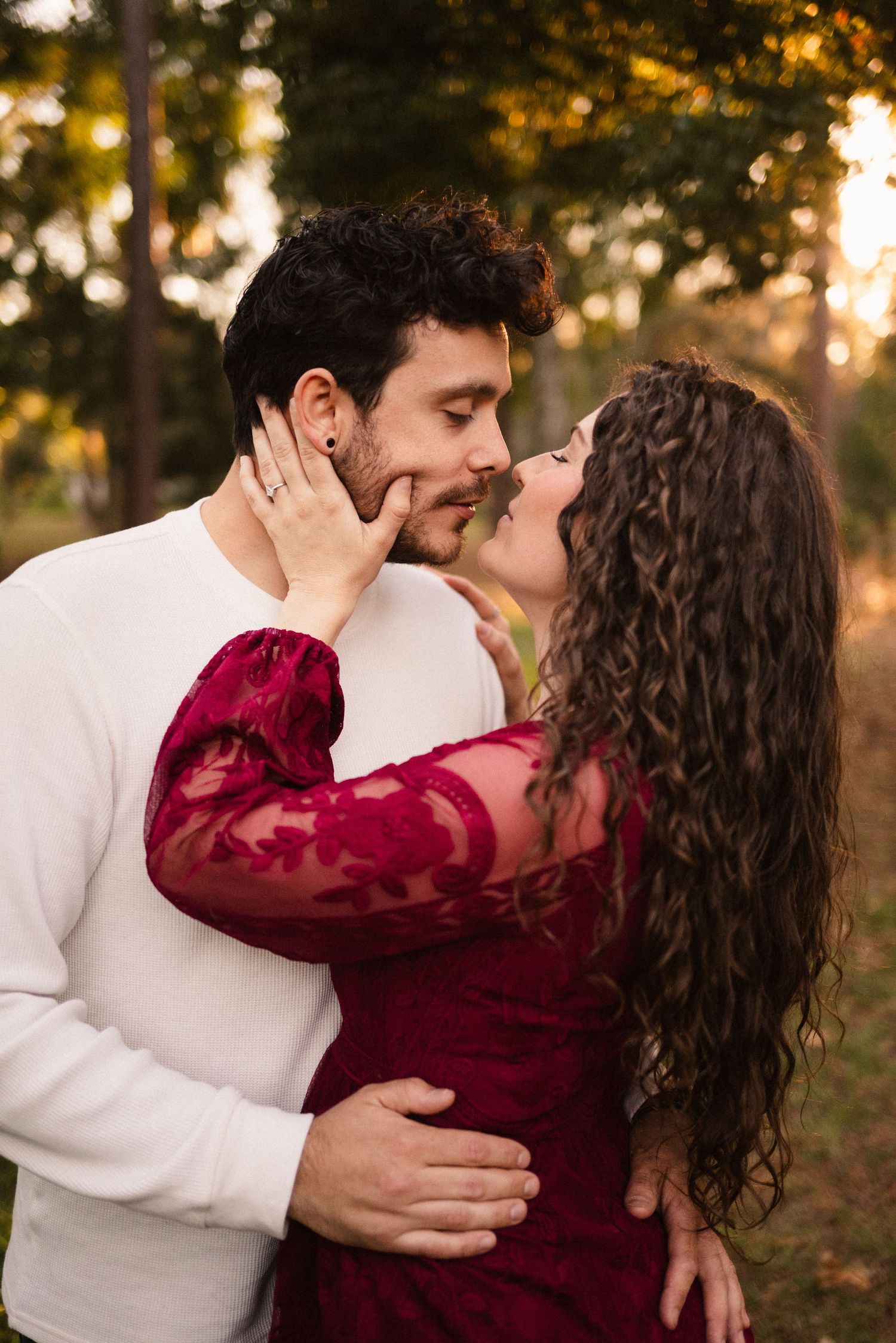 A couple shares an intimate moment in a park wearing burgundy and white during sunset.