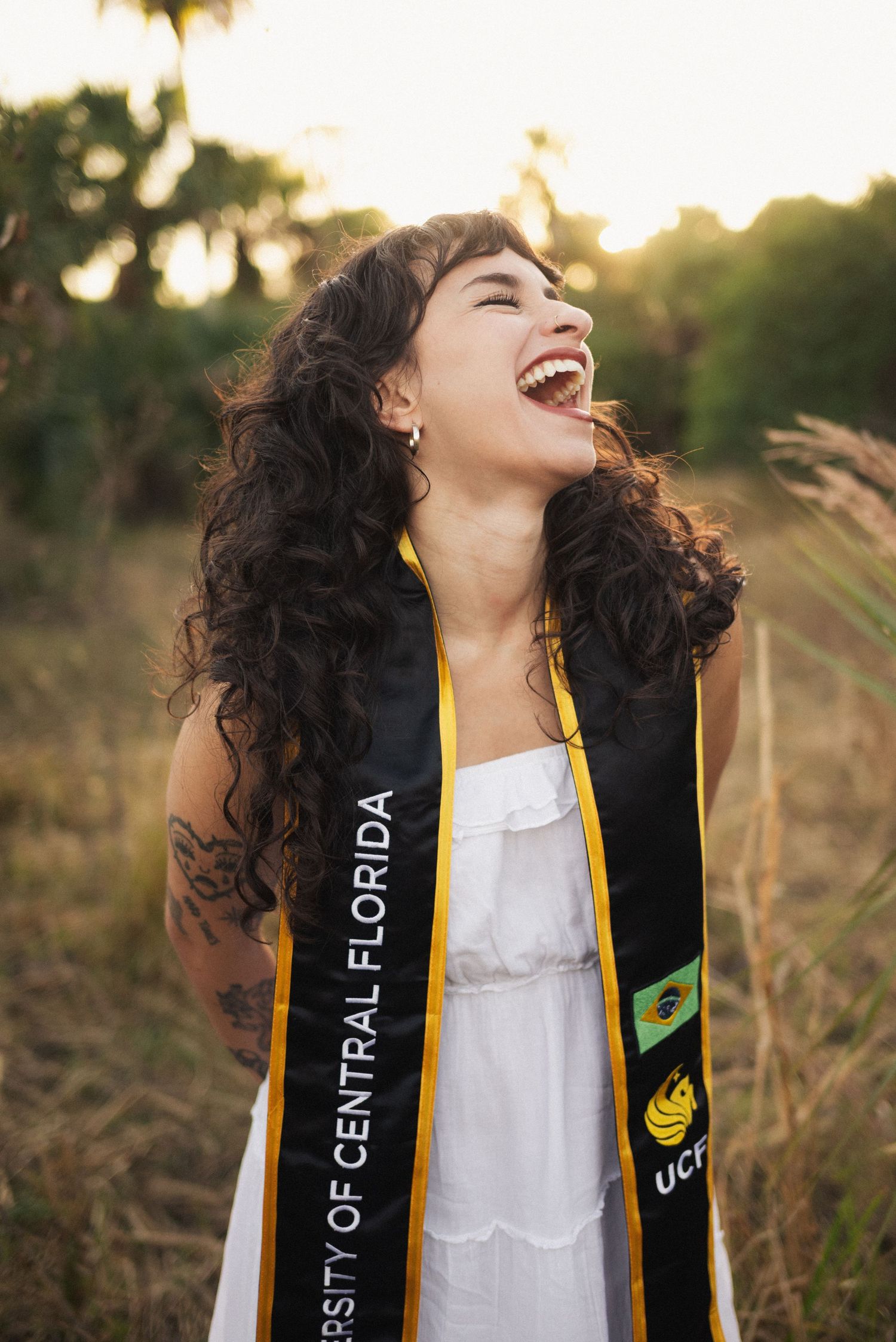 Graduate in white dress wearing academic stole laughs joyfully during sunset photo session.