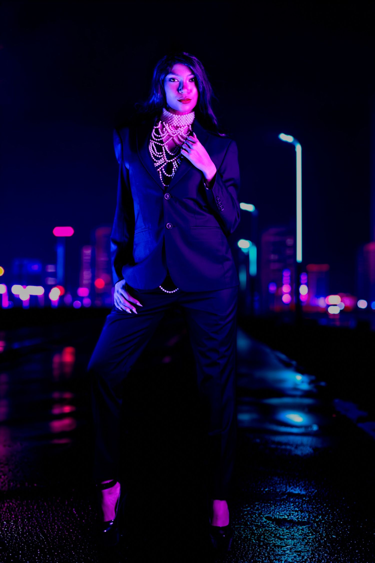 Woman in black suit with pearl necklace posing outdoors at night with colorful city bokeh lights in background.