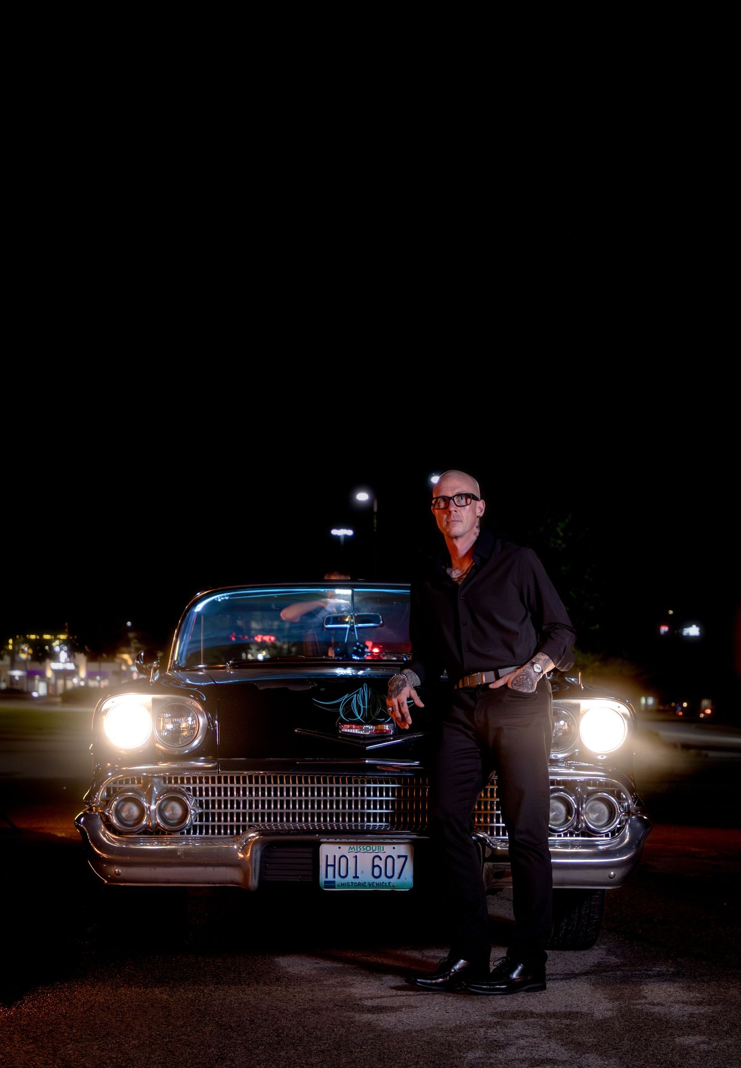 Man in black outfit leaning on a classic vintage black car at night with headlights on.