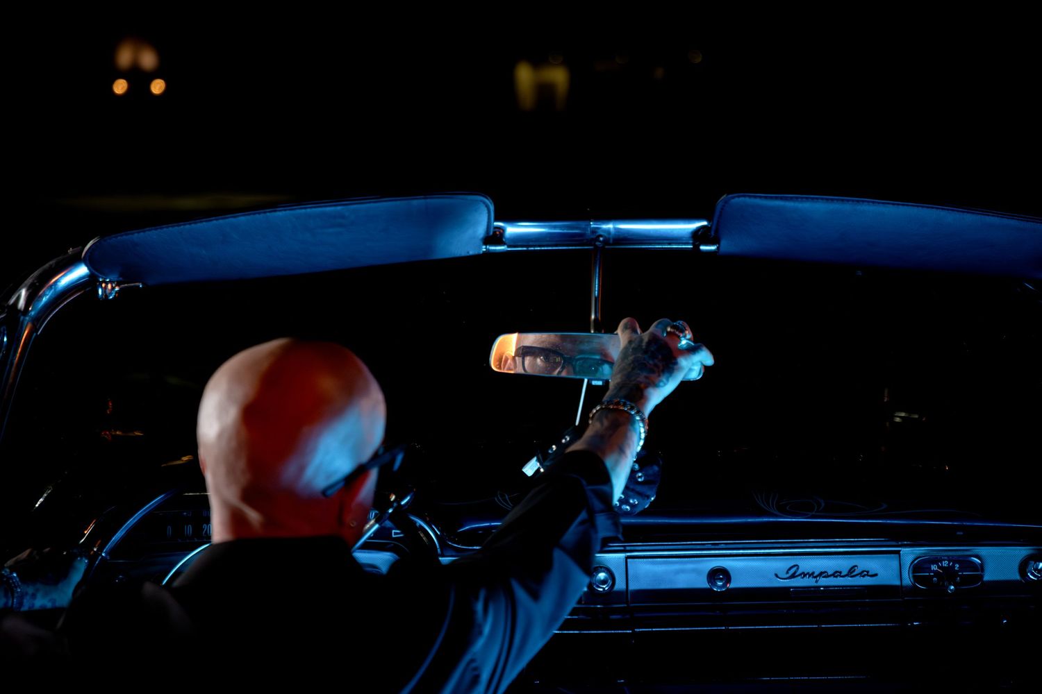 Bald man reaching up to adjust rearview mirror in classic Impala convertible at night.