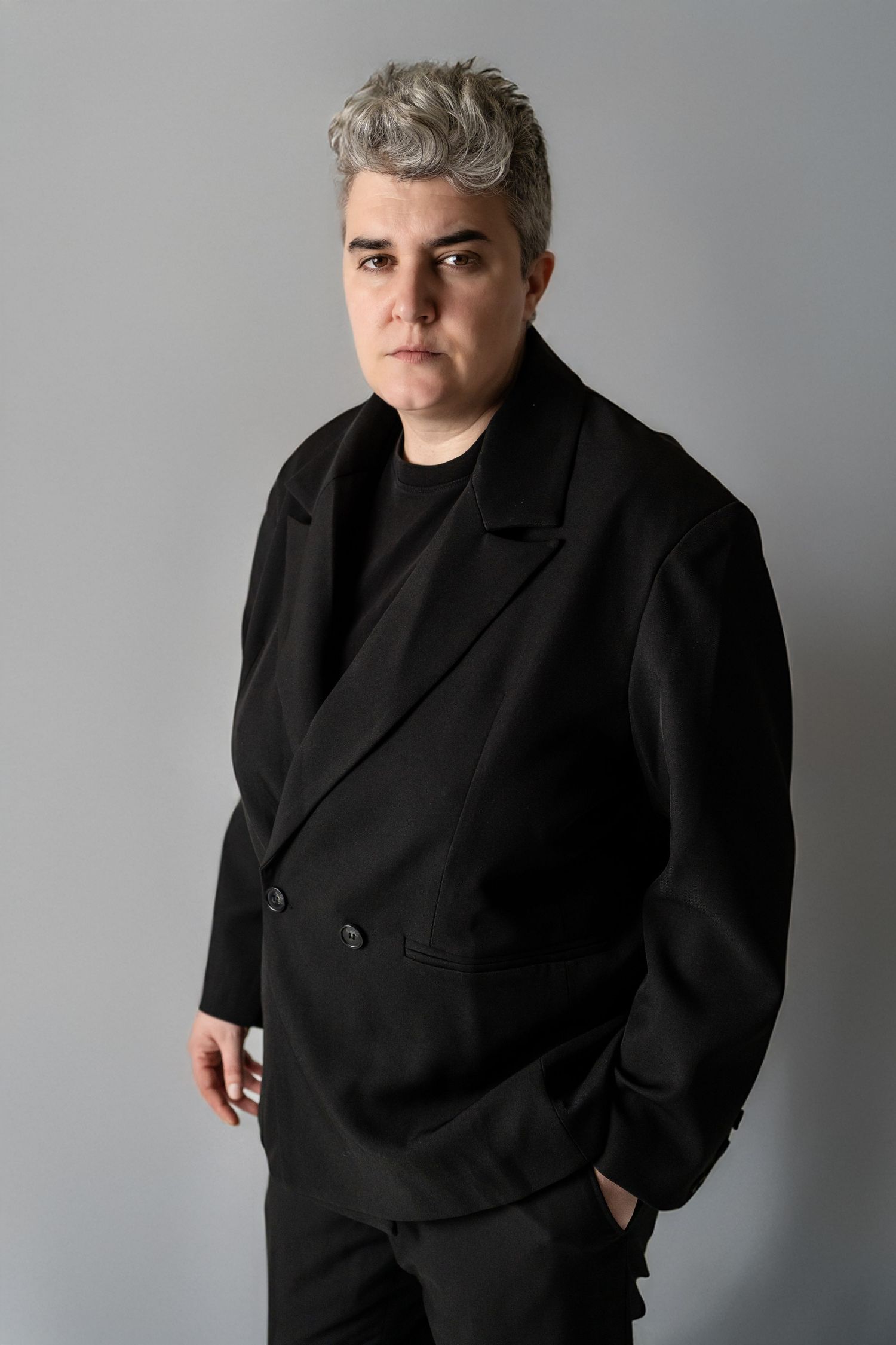 Person with short gray hair wearing an all-black blazer and pants, posing against a gray background.