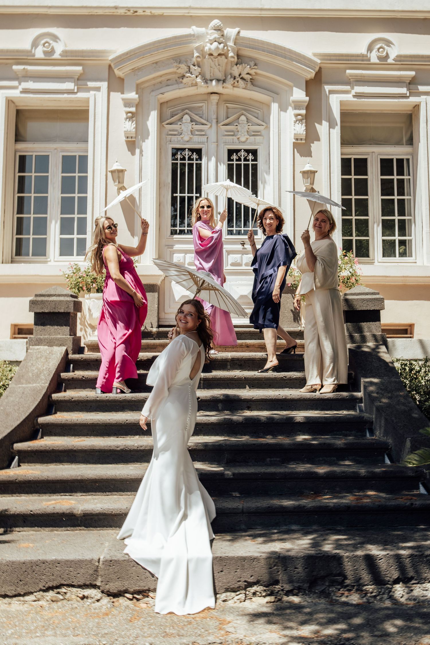 Bride in white gown poses with bridesmaids in colorful dresses on steps of elegant building.