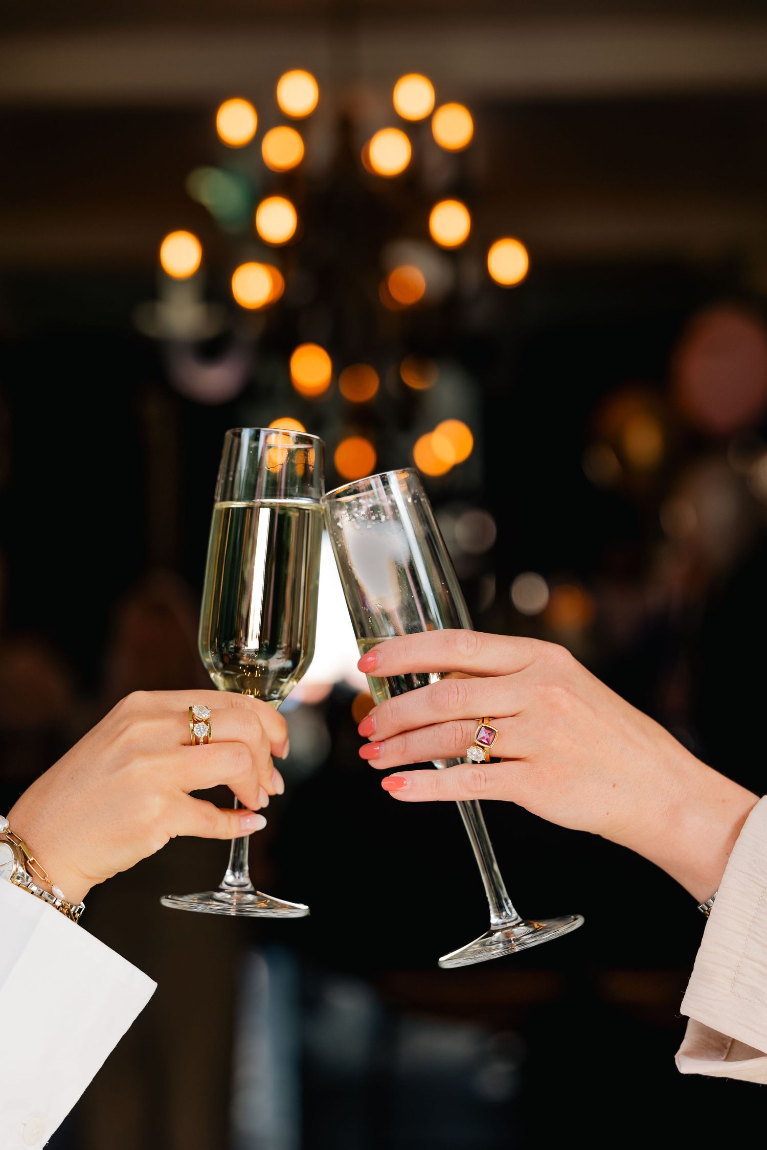 Two hands holding wine glasses clink together in a toast against a dark background with glowing bokeh lights.