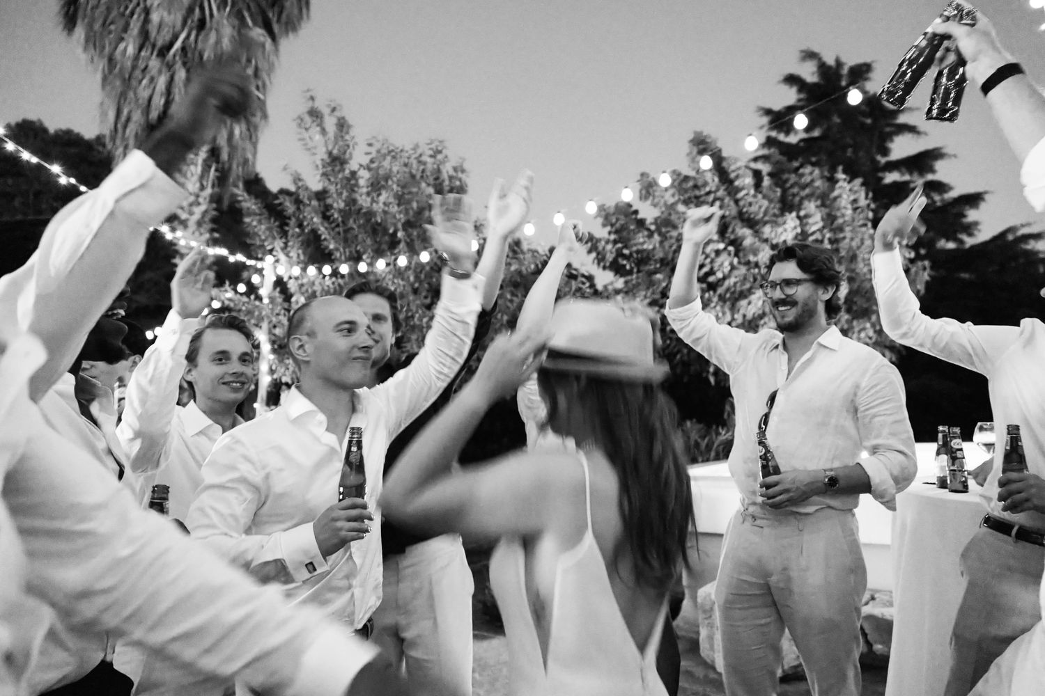 Guests dancing outdoors at a wedding reception with string lights and palm trees in black and white.