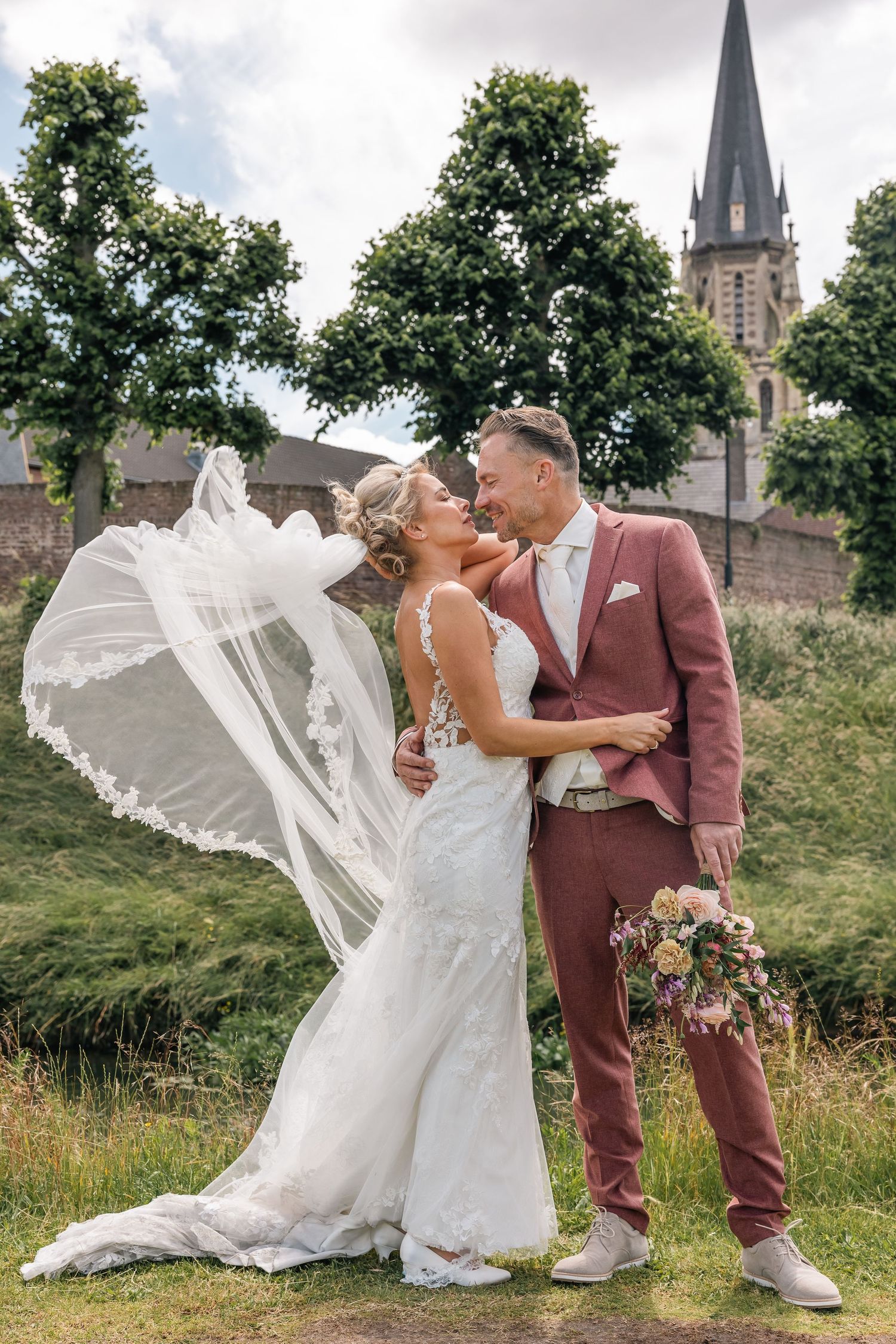 Newlyweds share a romantic kiss in front of a church spire with bride's veil flowing in the wind.