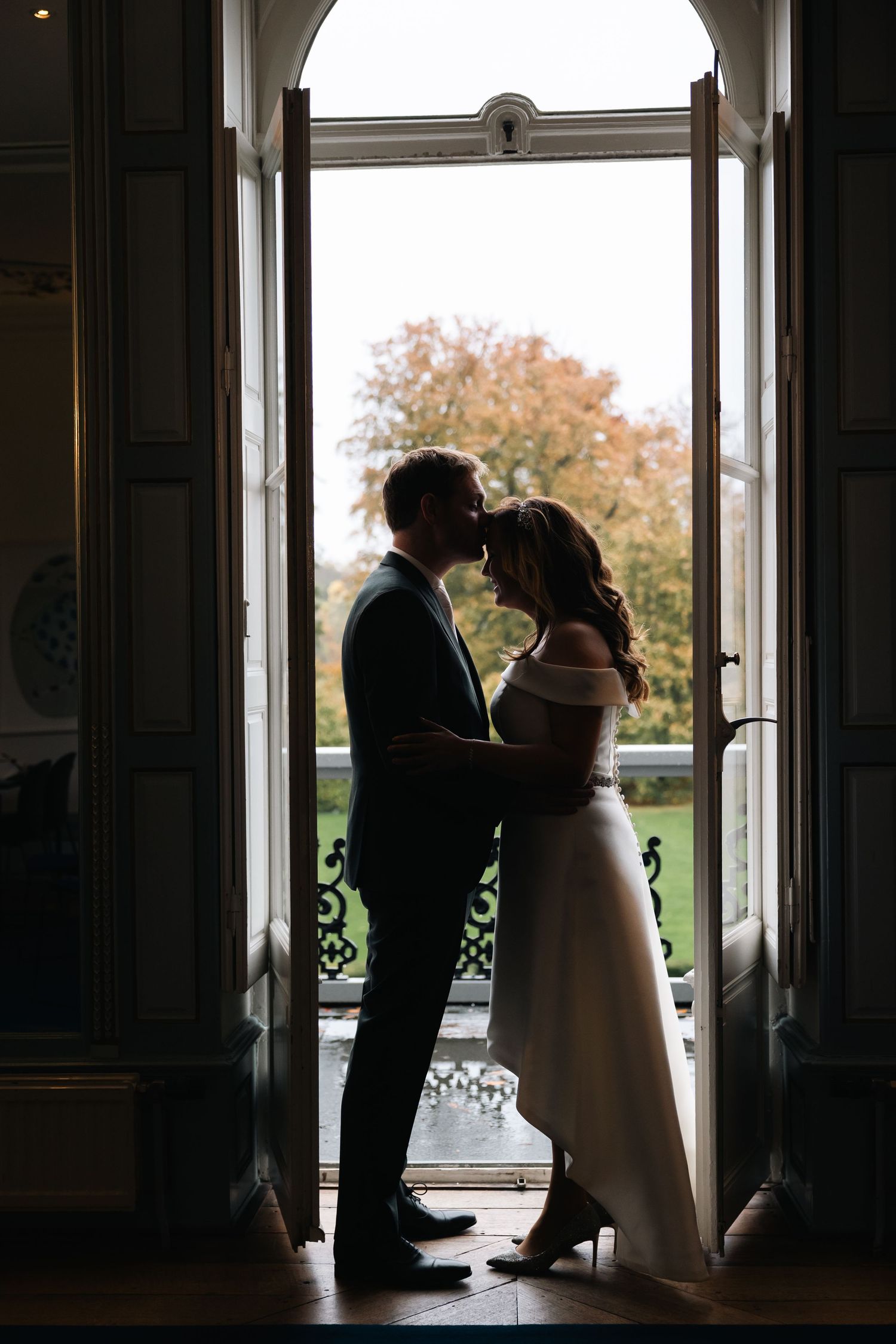 Silhouetted couple shares romantic moments by large windows in an elegant indoor setting during their wedding day.