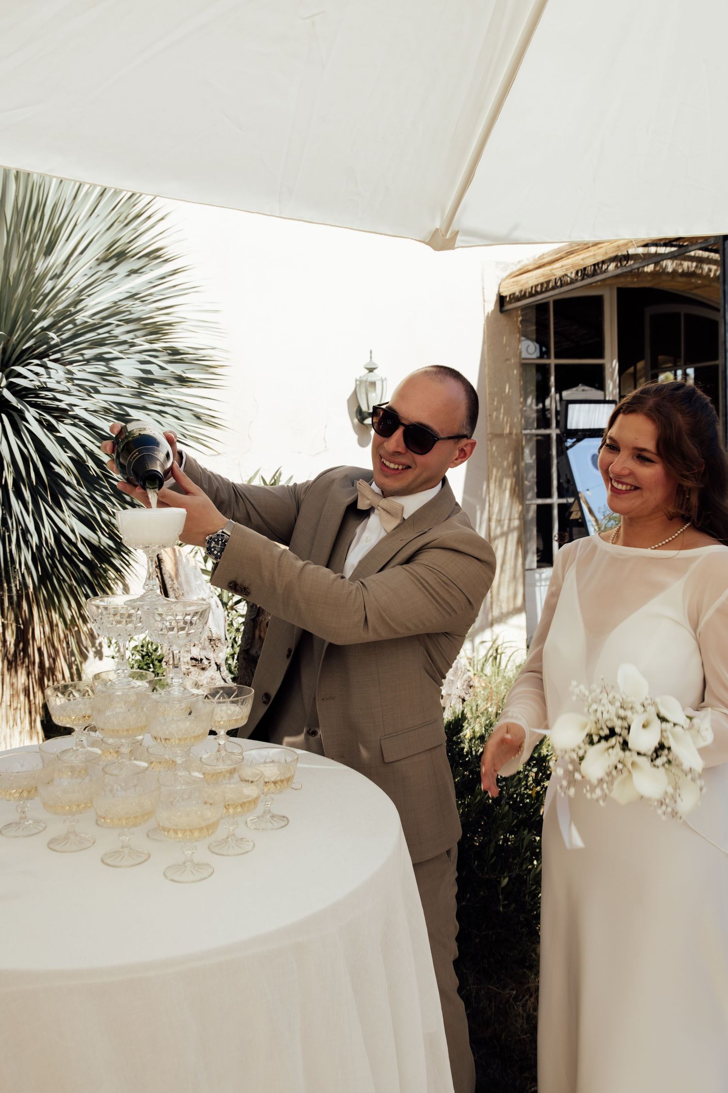 Groom in tan suit pouring champagne tower at wedding reception while bride in white gown holds bouquet.
