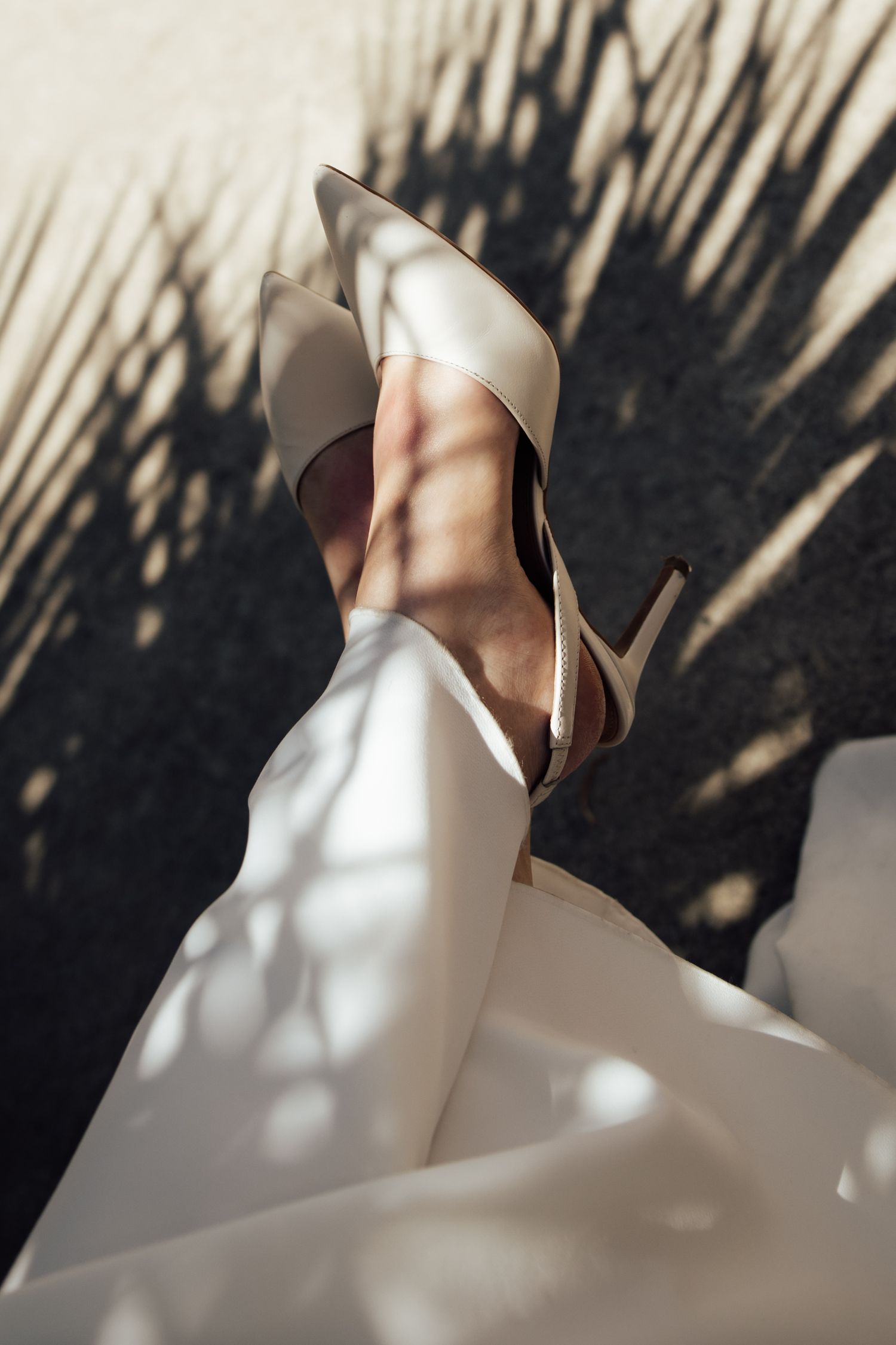 A woman's leg raised wearing elegant ivory satin pointed-toe stiletto heels against a sunlit wall with palm leaf shadows.