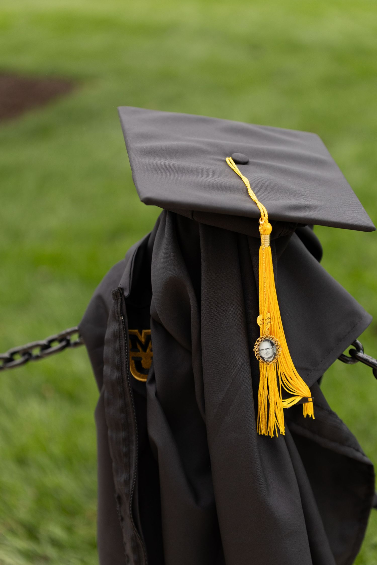 Black graduation cap and gown with gold tassel draped over iron chain fence on green lawn.