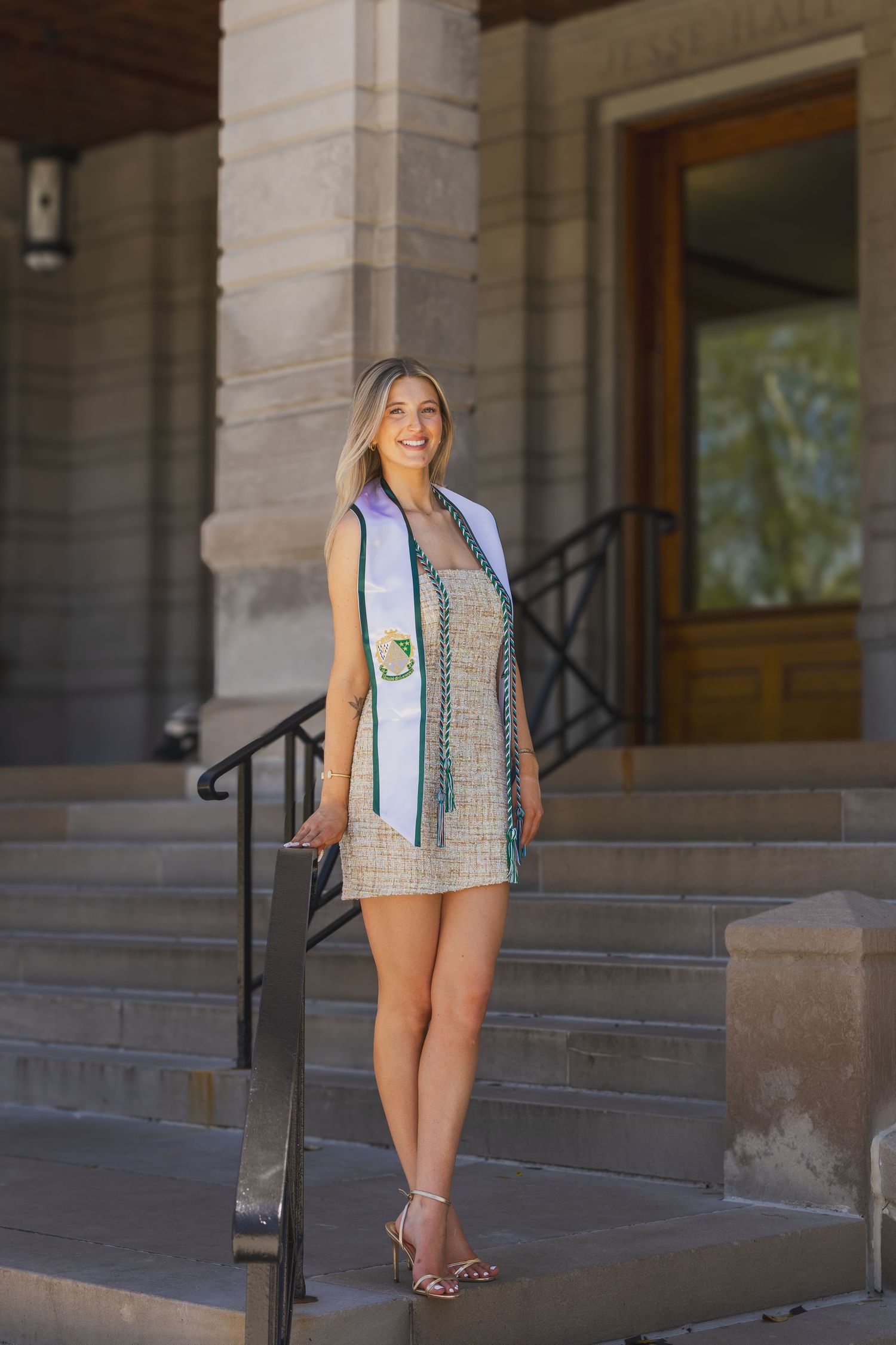 Smiling female graduate in beige dress with white and green honor stole on stone building steps.