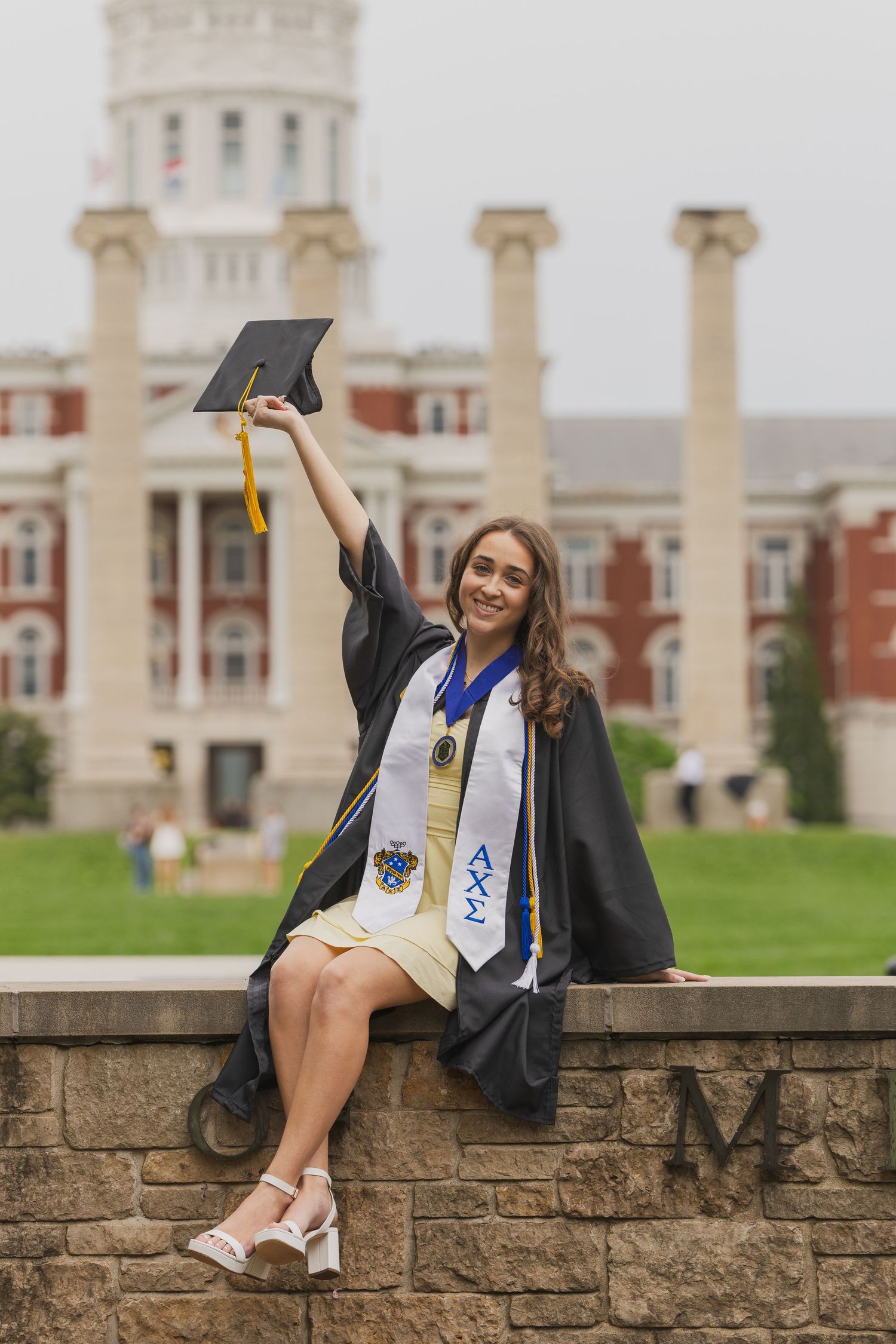 Happy female graduate raises mortarboard cap while sitting on stone wall before university columns.