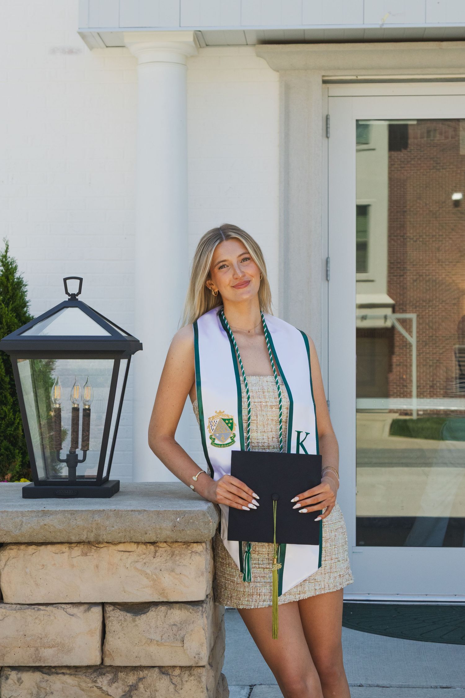 Female graduate standing by stone lantern holding diploma folder wearing white KD sorority graduation stole.