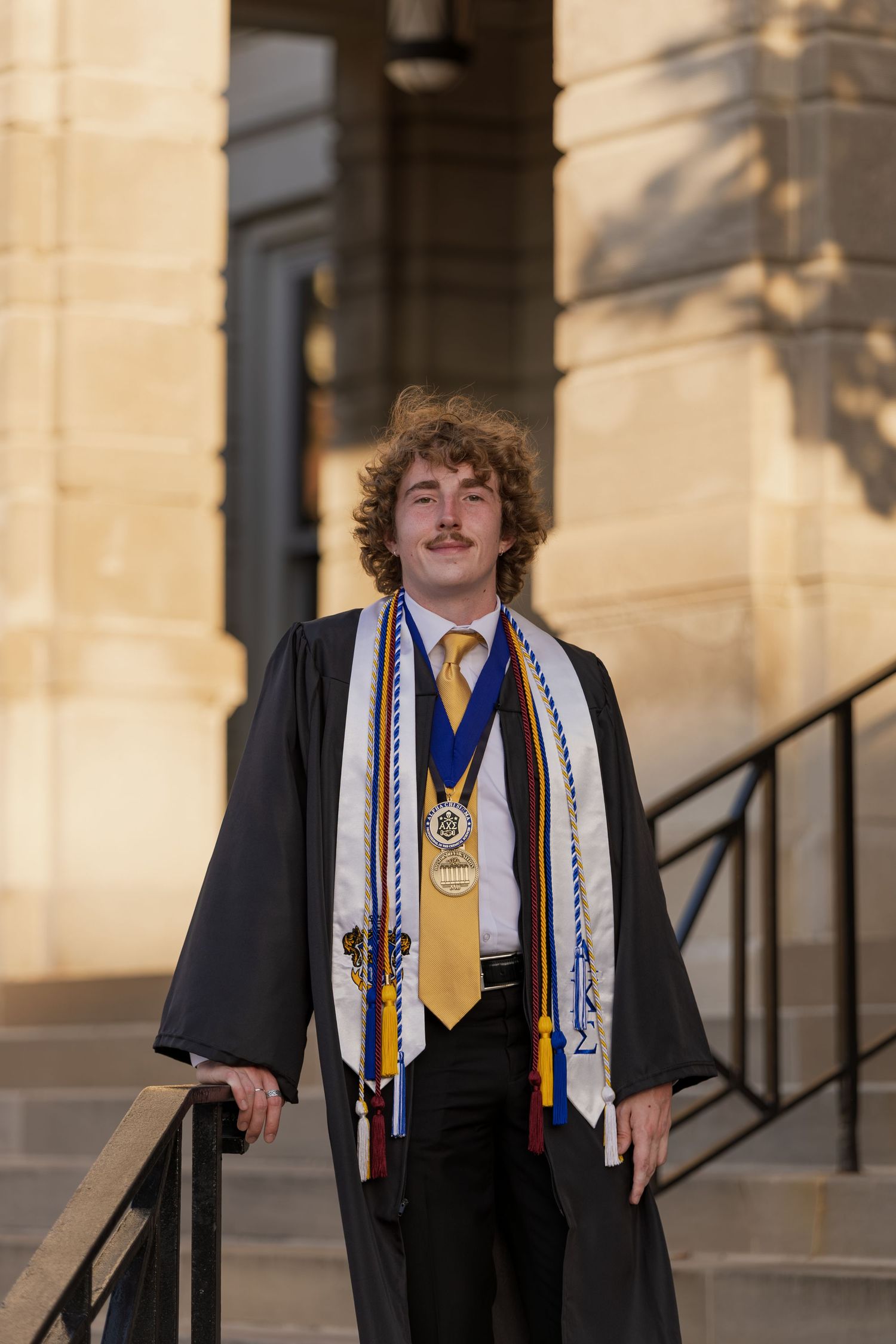 Male graduate with curly hair wearing black gown and multiple honor cords stands on steps.