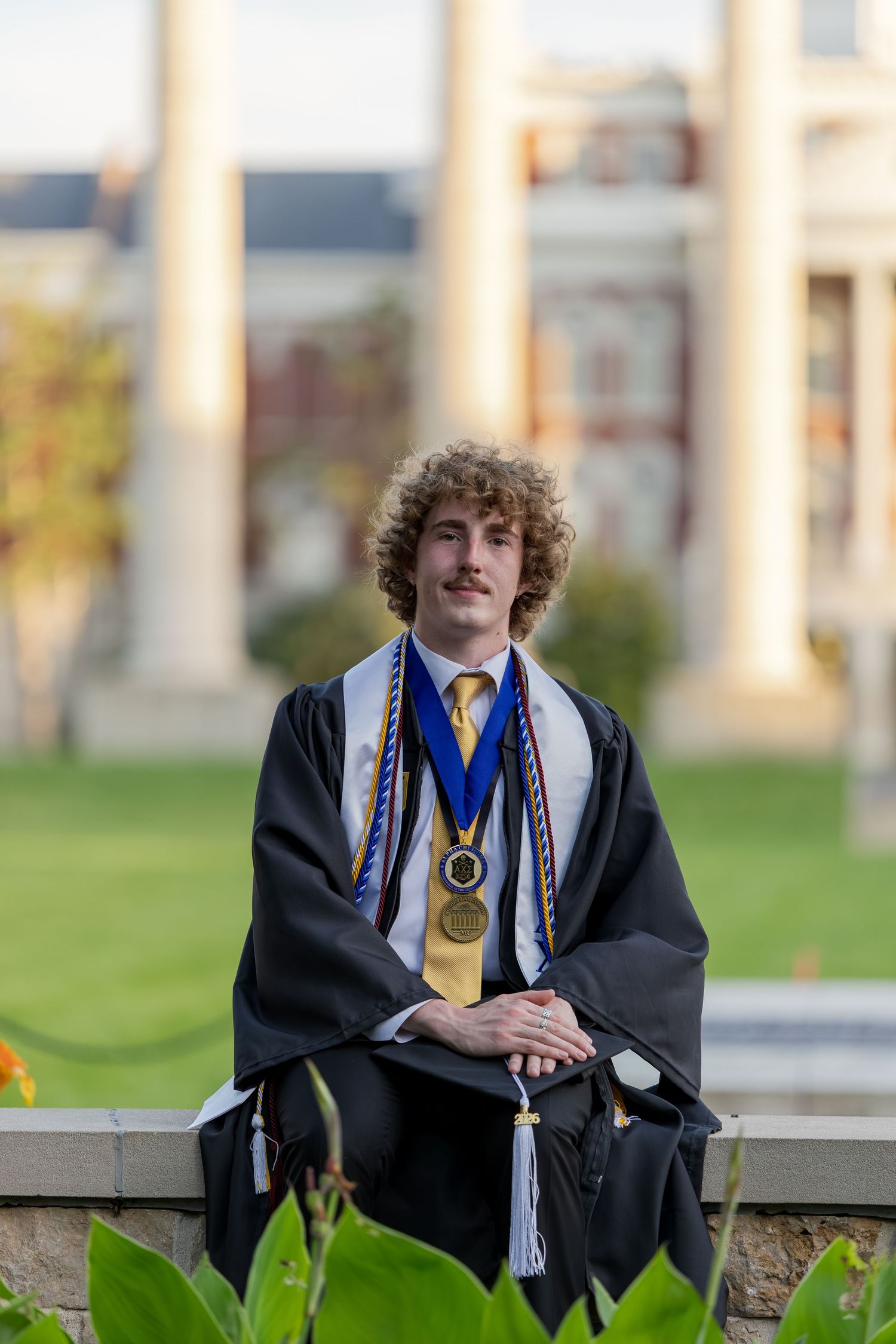 Male graduate in black gown with gold medal and honor cords sits before university columns.