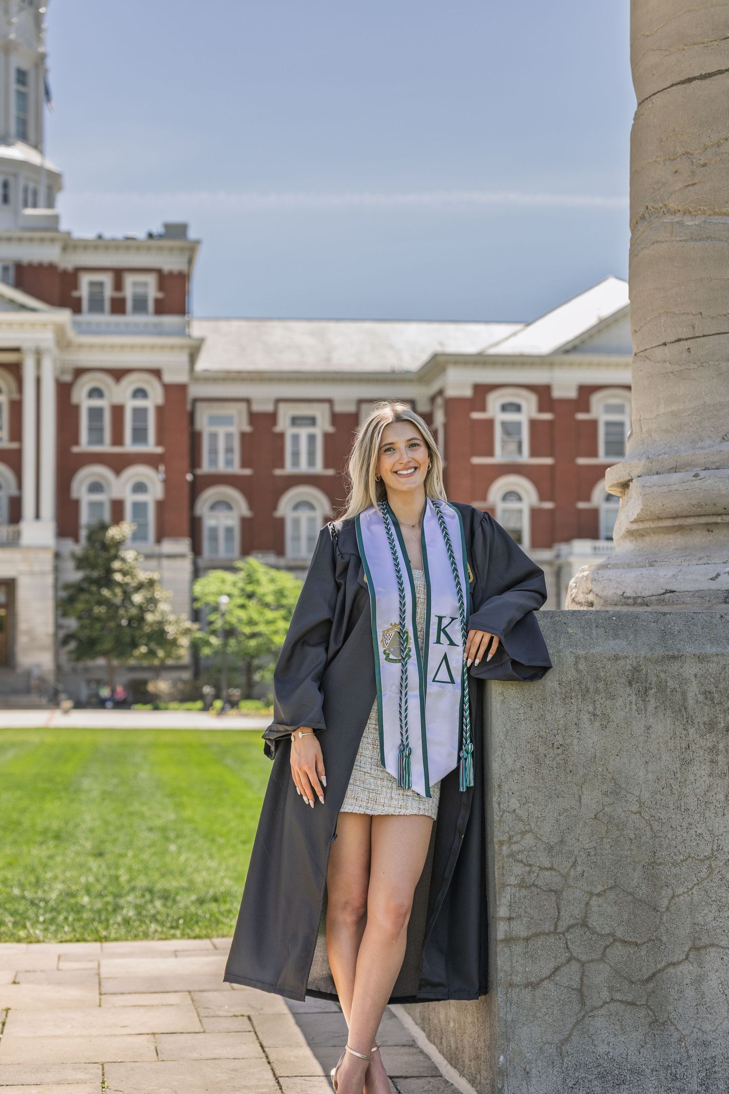 Female graduate in black gown leaning on column with brick university building in background.