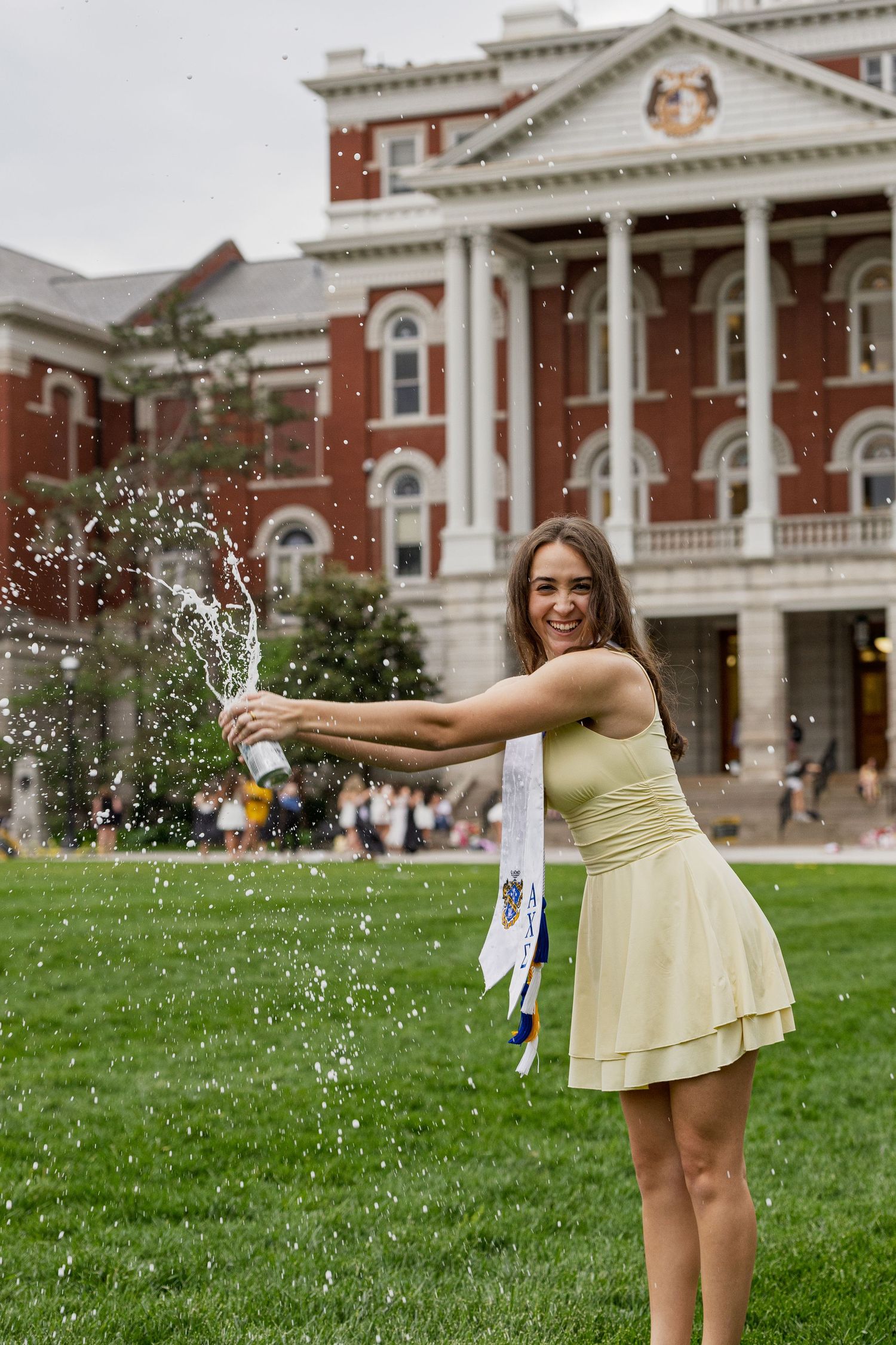 Joyful female graduate in yellow dress pops champagne bottle on green university campus lawn.
