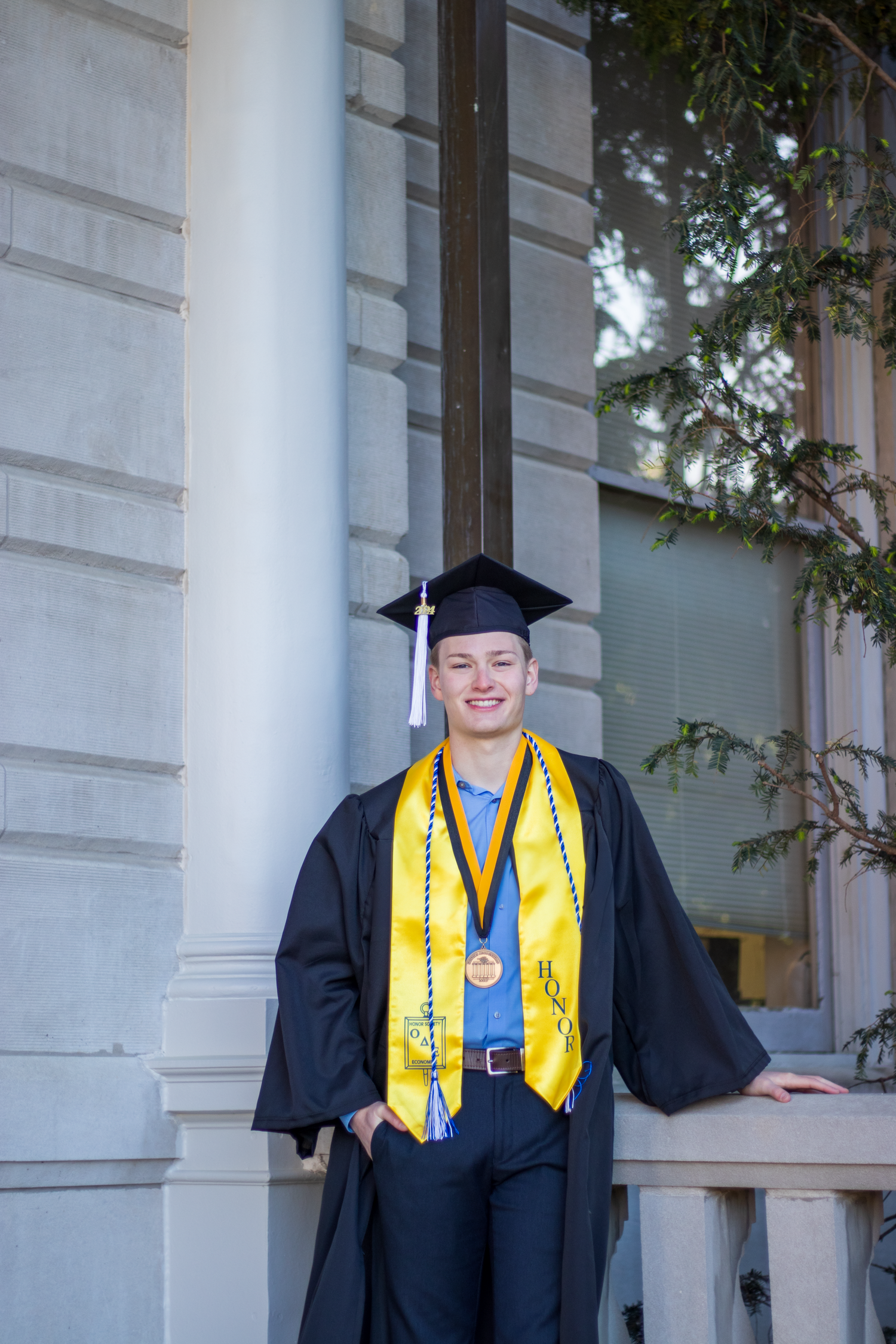 Graduate in black cap and gown with gold honor stole standing by white pillars outdoors.