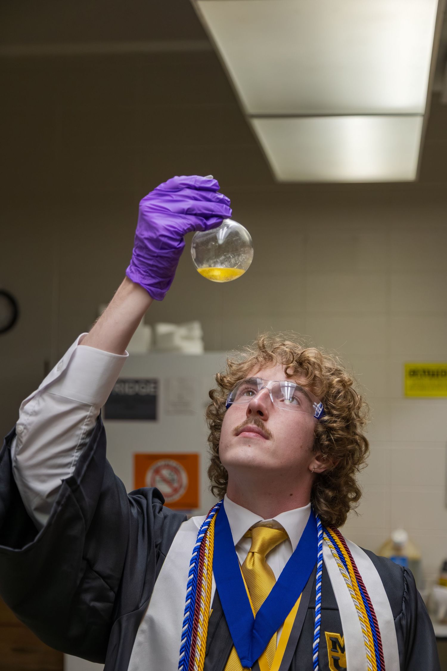 Graduate in black gown examining yellow liquid in flask while wearing purple gloves and safety goggles.