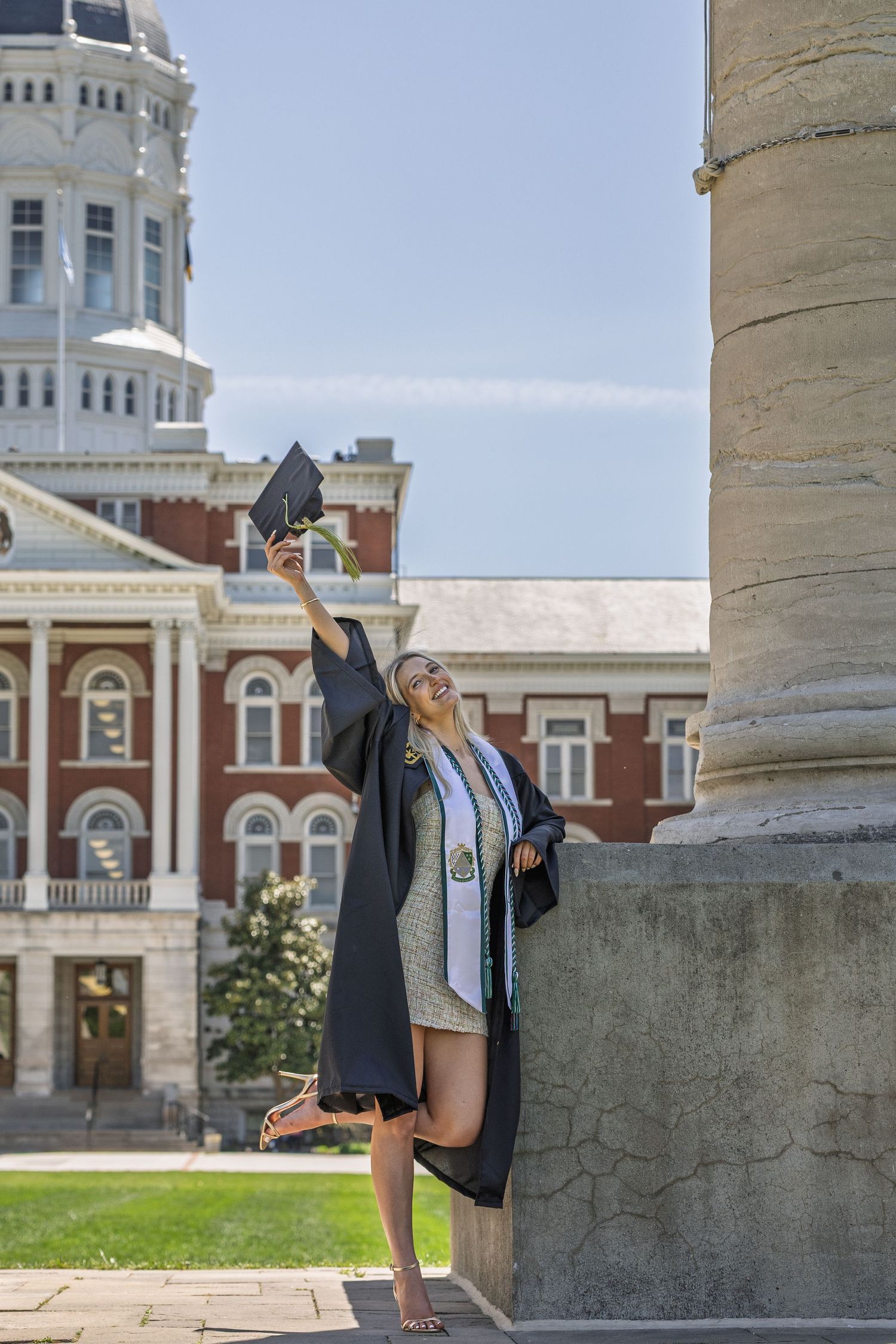 Female graduate in black gown raising cap beside stone column with ornate brick building behind her.