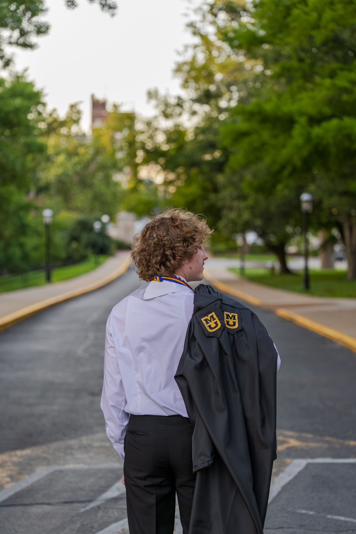 Male graduate carries black gown over shoulder while walking tree-lined university campus path.