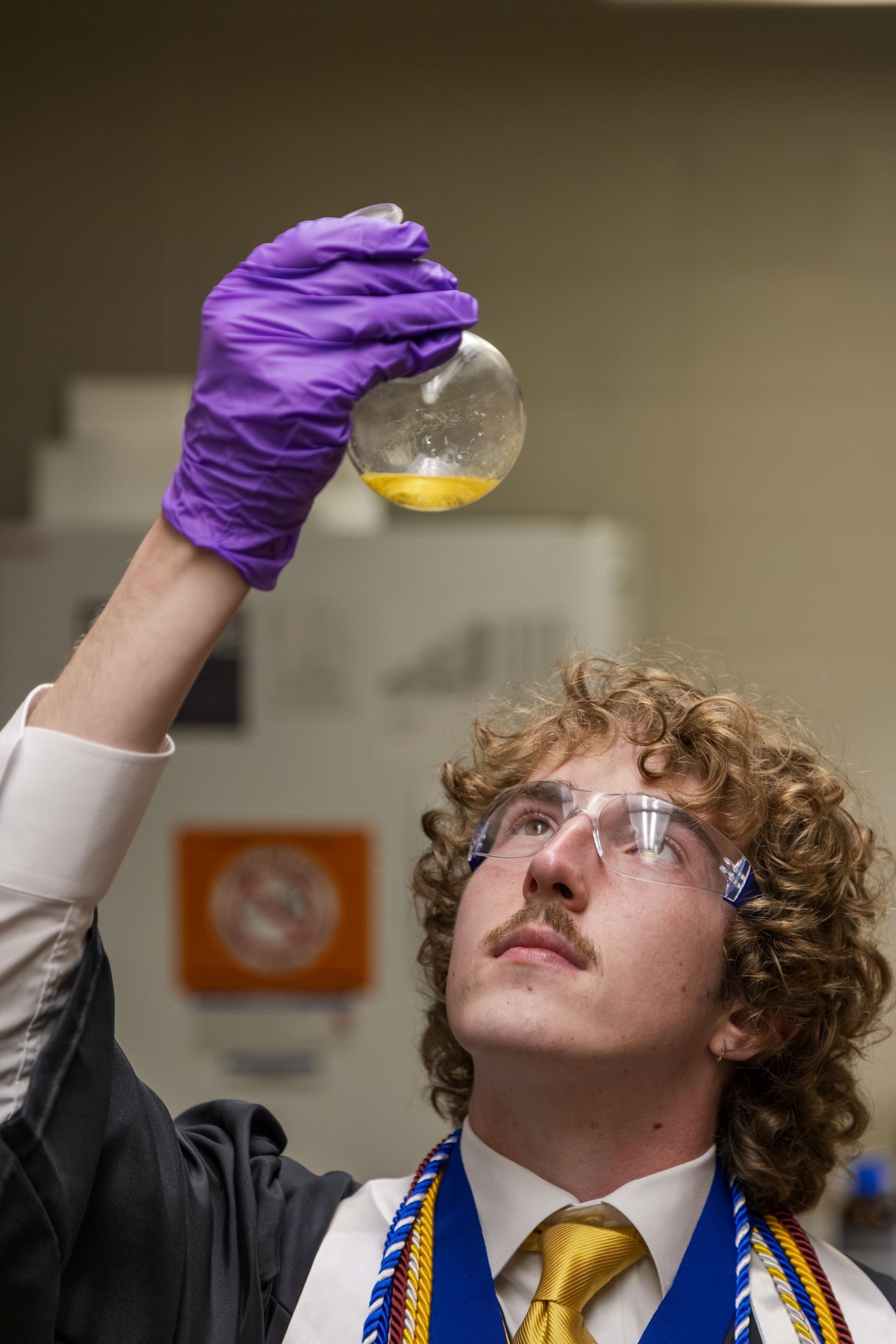 Male graduate in lab coat examines flask with yellow liquid while wearing safety goggles.