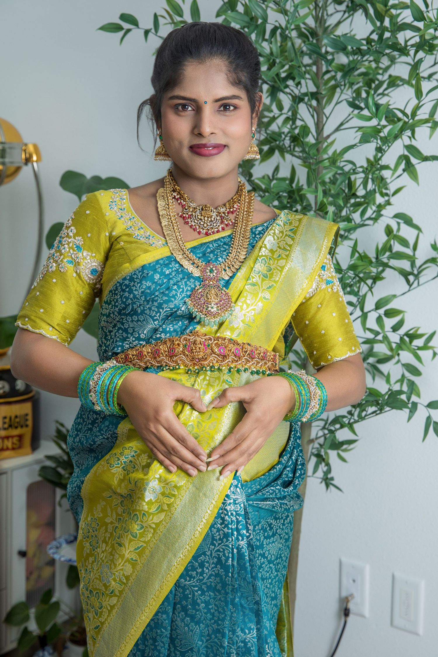 Woman in teal and yellow silk saree with gold jewelry making heart shape gesture over baby bump.