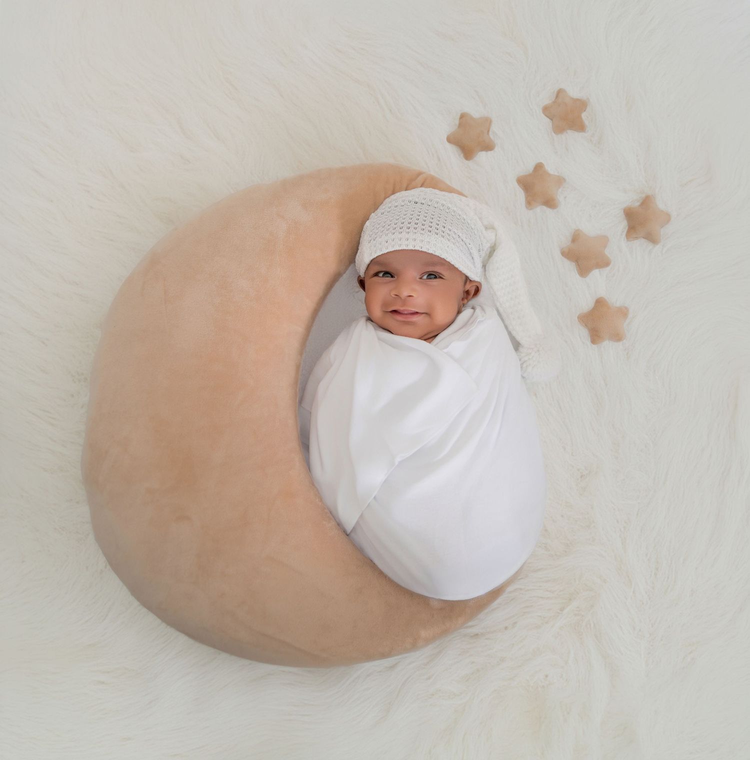 Newborn baby swaddled in white on a crescent moon pillow with wooden stars on a fluffy white rug.
