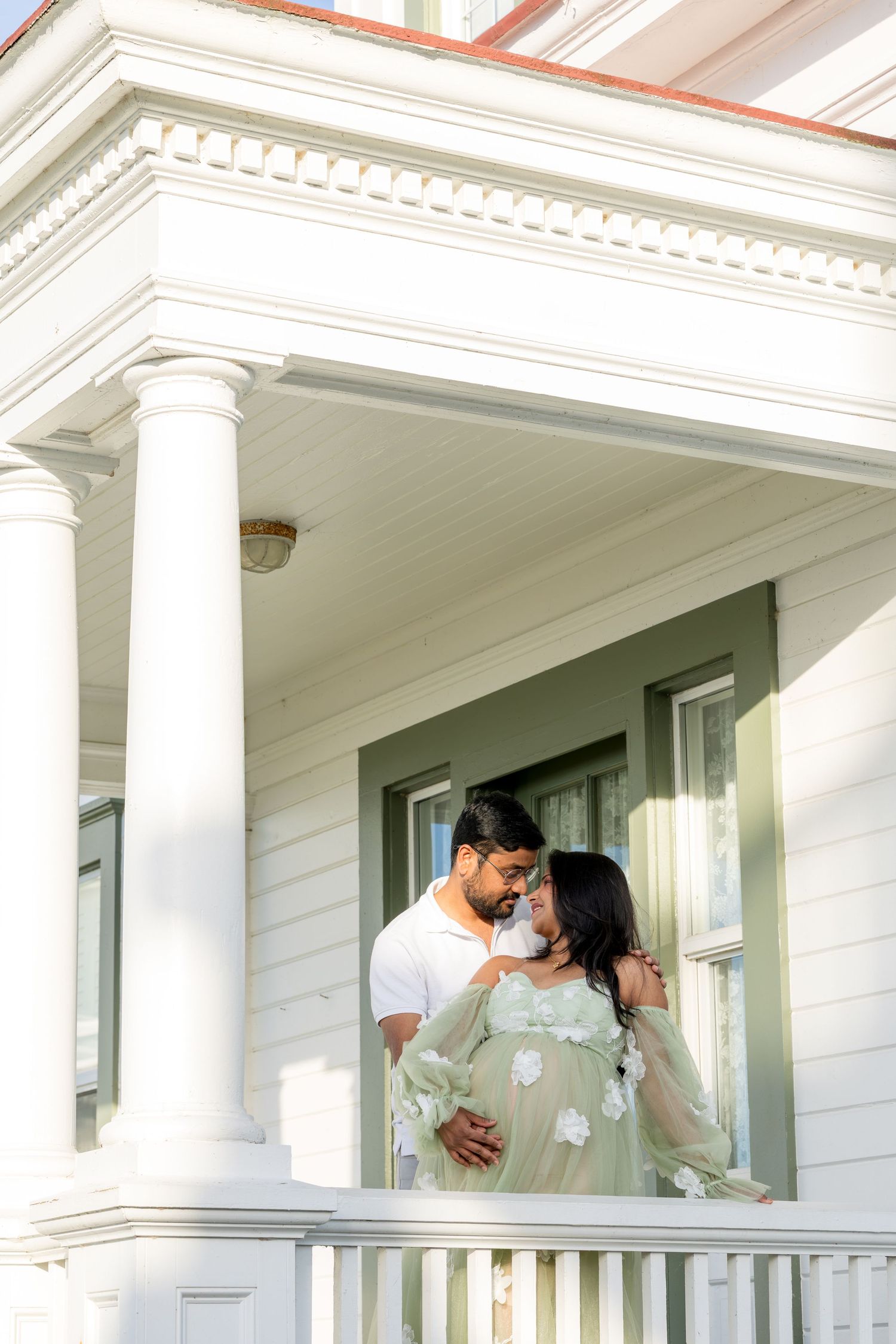 Couple sharing a tender moment on a white colonial porch, woman in sage green maternity gown, sunny day.
