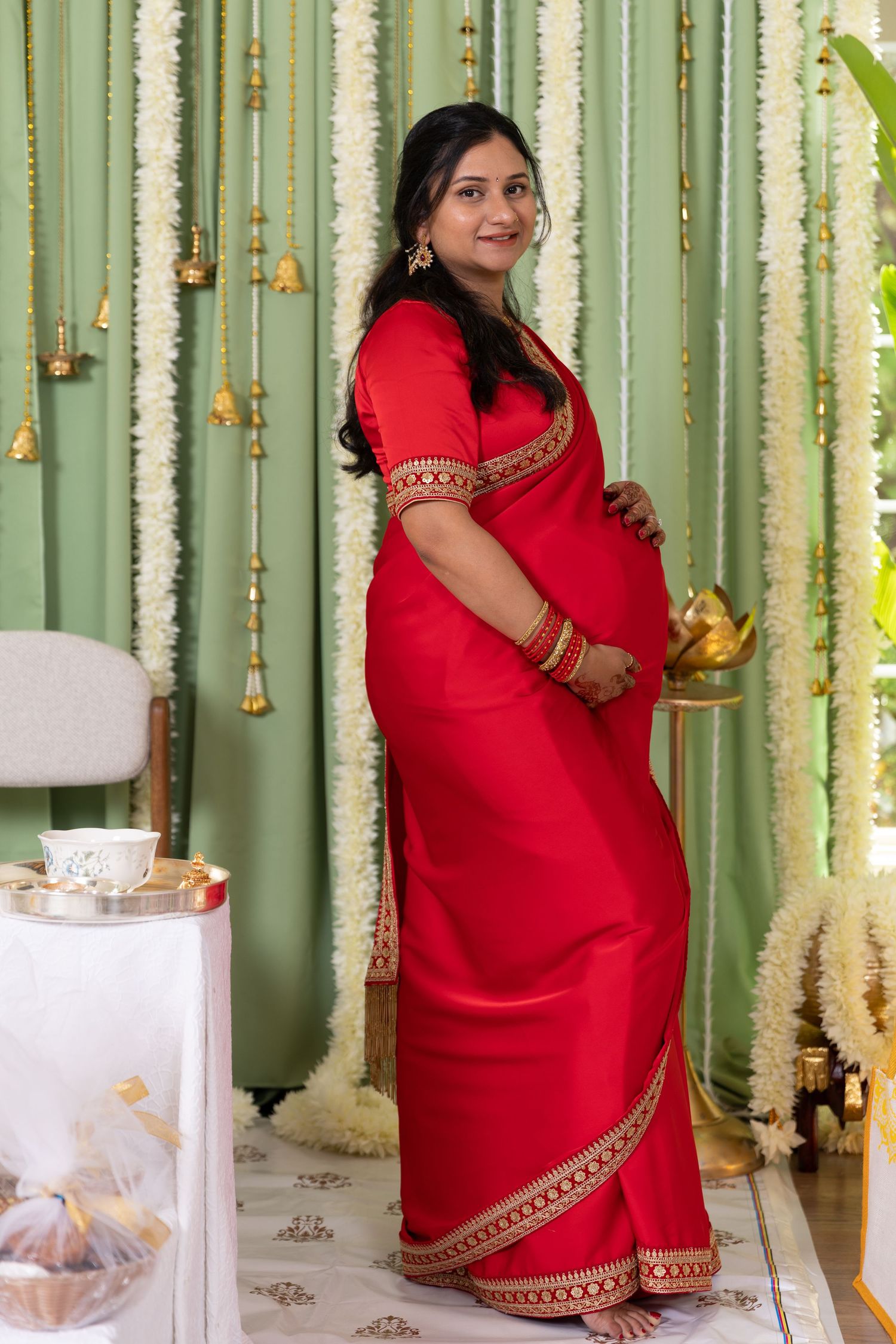 Pregnant woman in red saree with gold trim poses at traditional Indian baby shower ceremony with floral decorations.