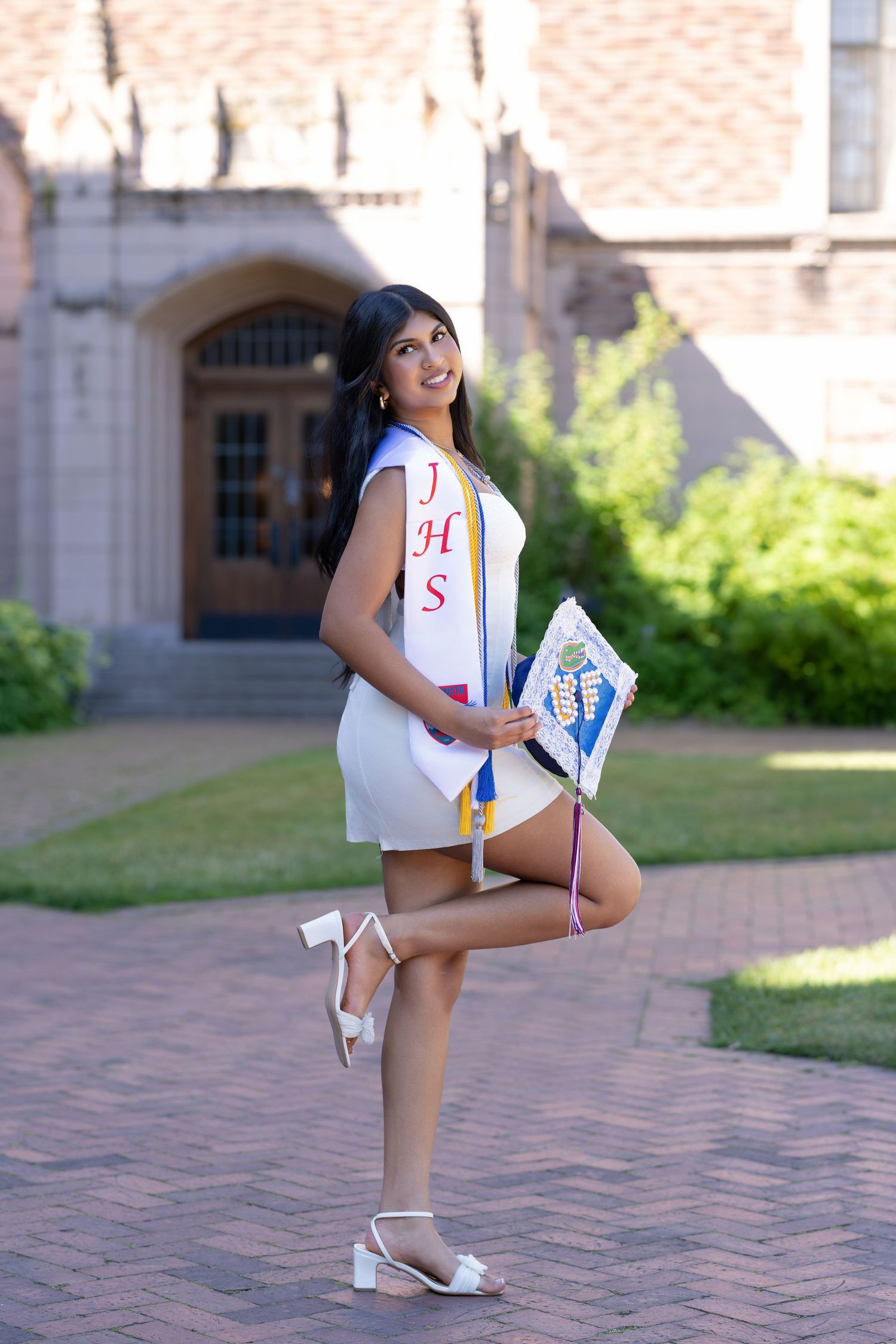 A young woman in a white dress and JHS sash poses with a decorated graduation cap on a brick path.