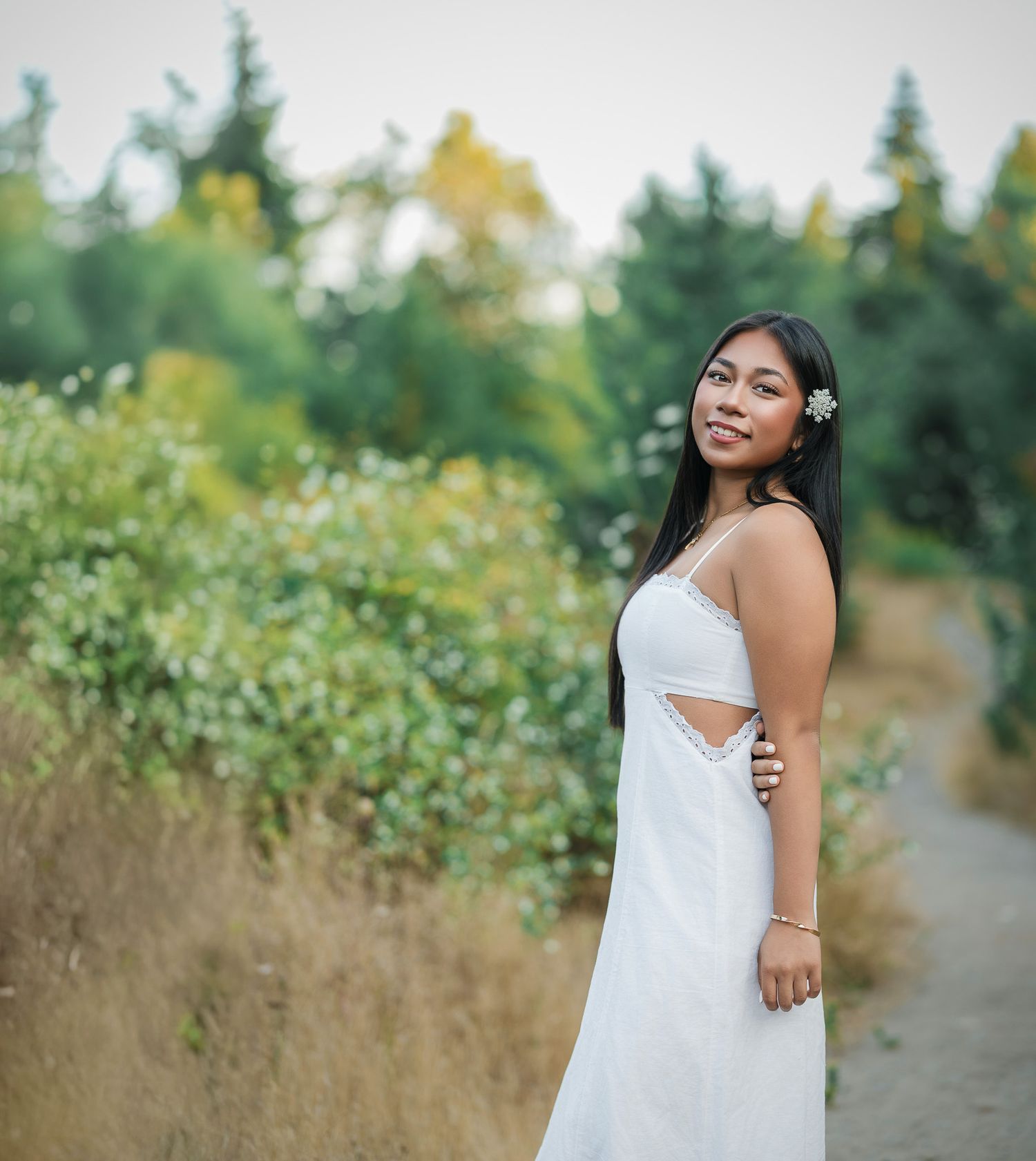 Young woman in white sundress smiling on a garden path surrounded by lush green trees and shrubs.