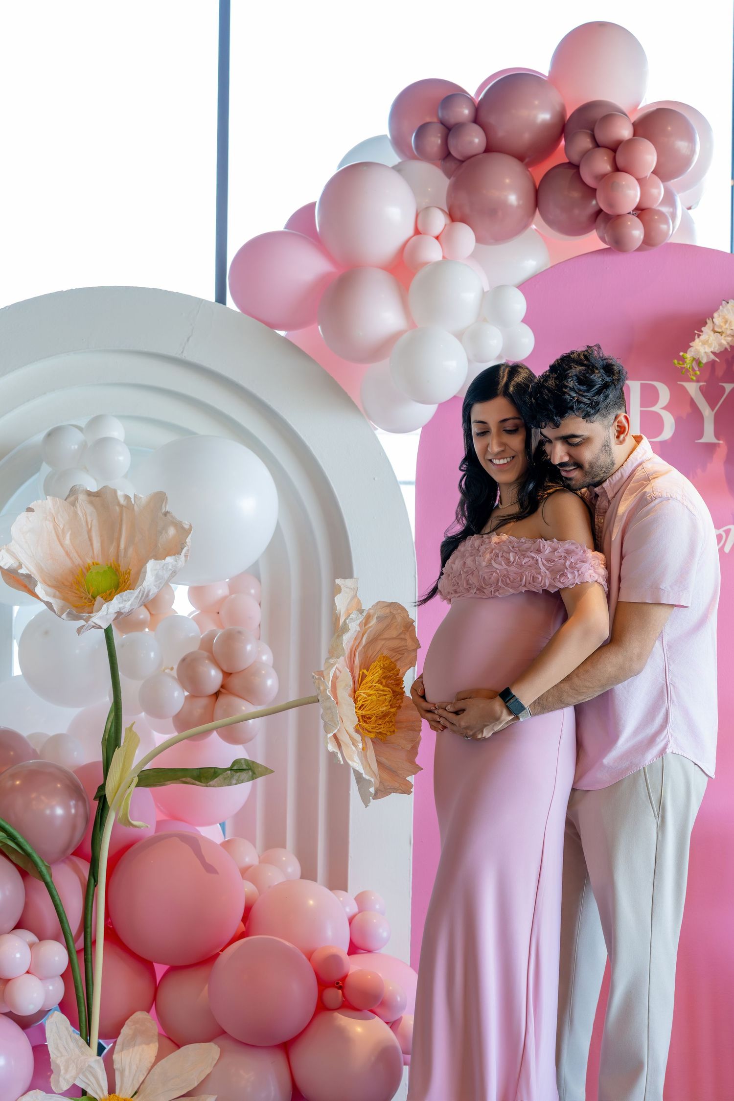 Couple at a pink-themed baby shower, surrounded by balloon garlands and floral decor, cradling a baby bump.
