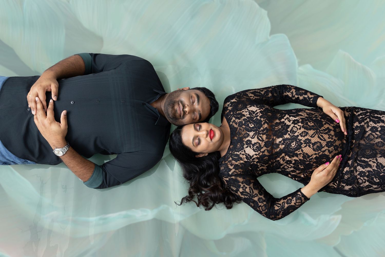 A couple lies on teal fabric, man in blue suit and woman in black lace maternity gown, heads together.