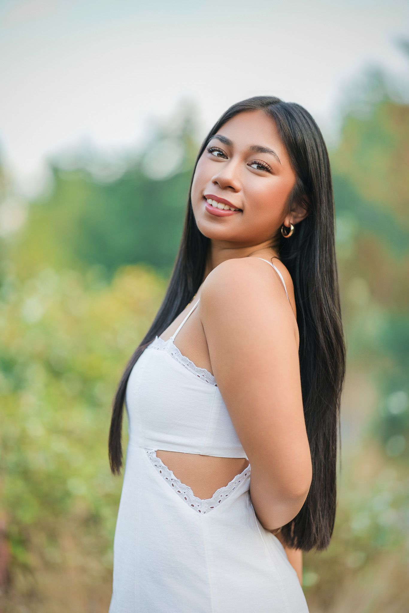 Young woman in white sundress with long black hair smiling outdoors with green trees in background.
