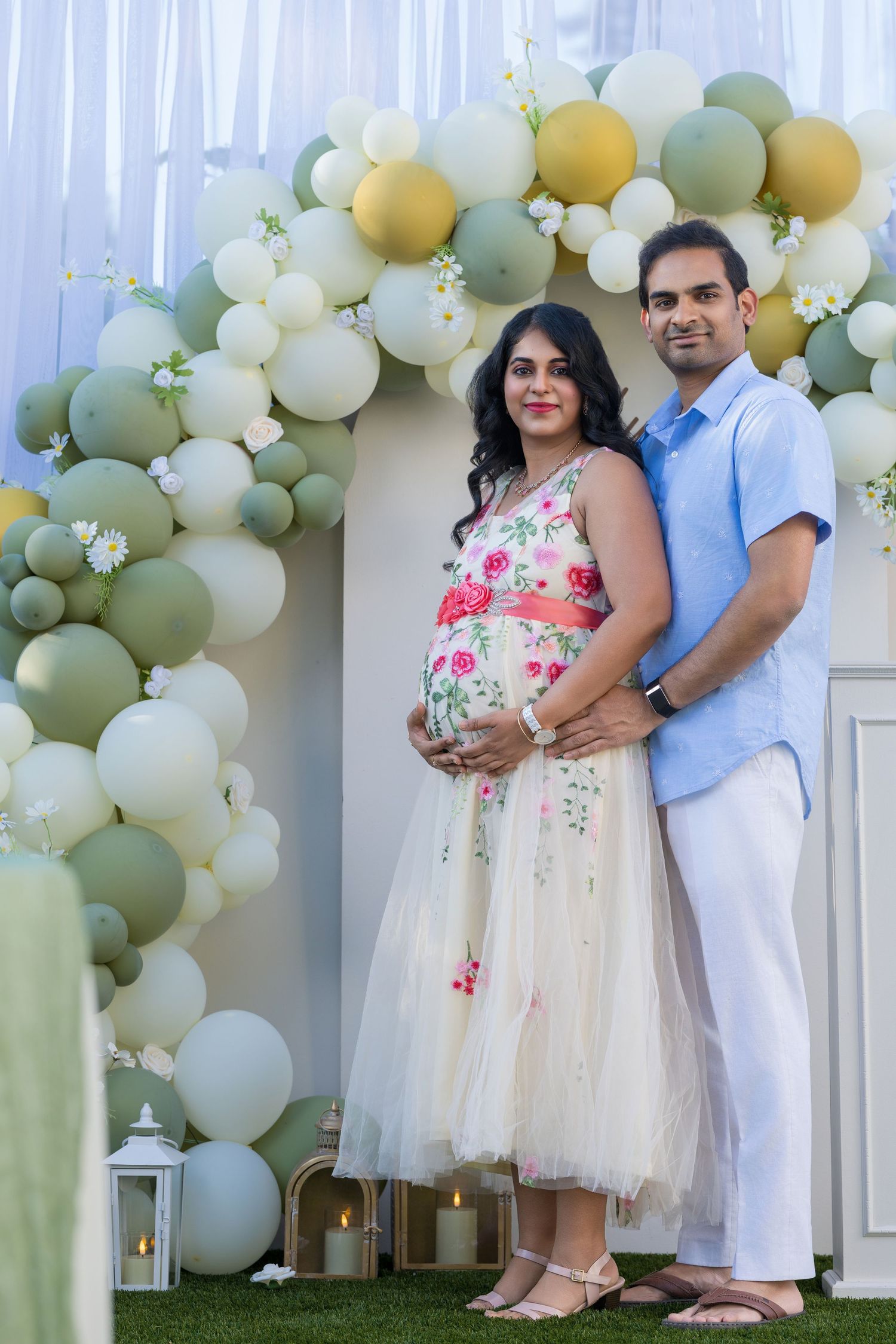 Pregnant couple posing under green and white balloon arch at baby shower celebration outdoors.