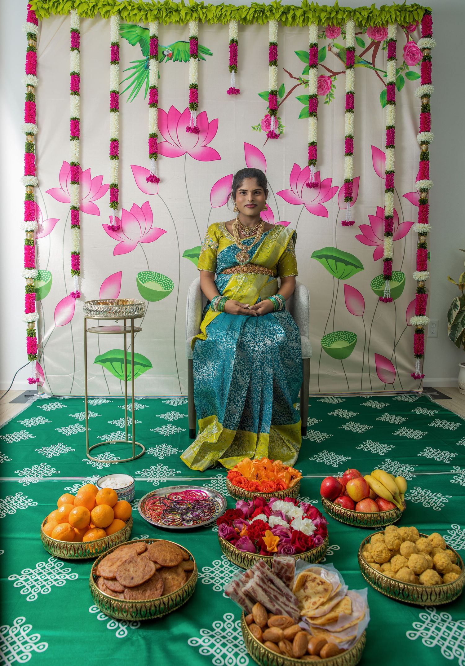 Woman in colorful saree sits before offerings of fruit, sweets, and flowers on green floor with floral backdrop.